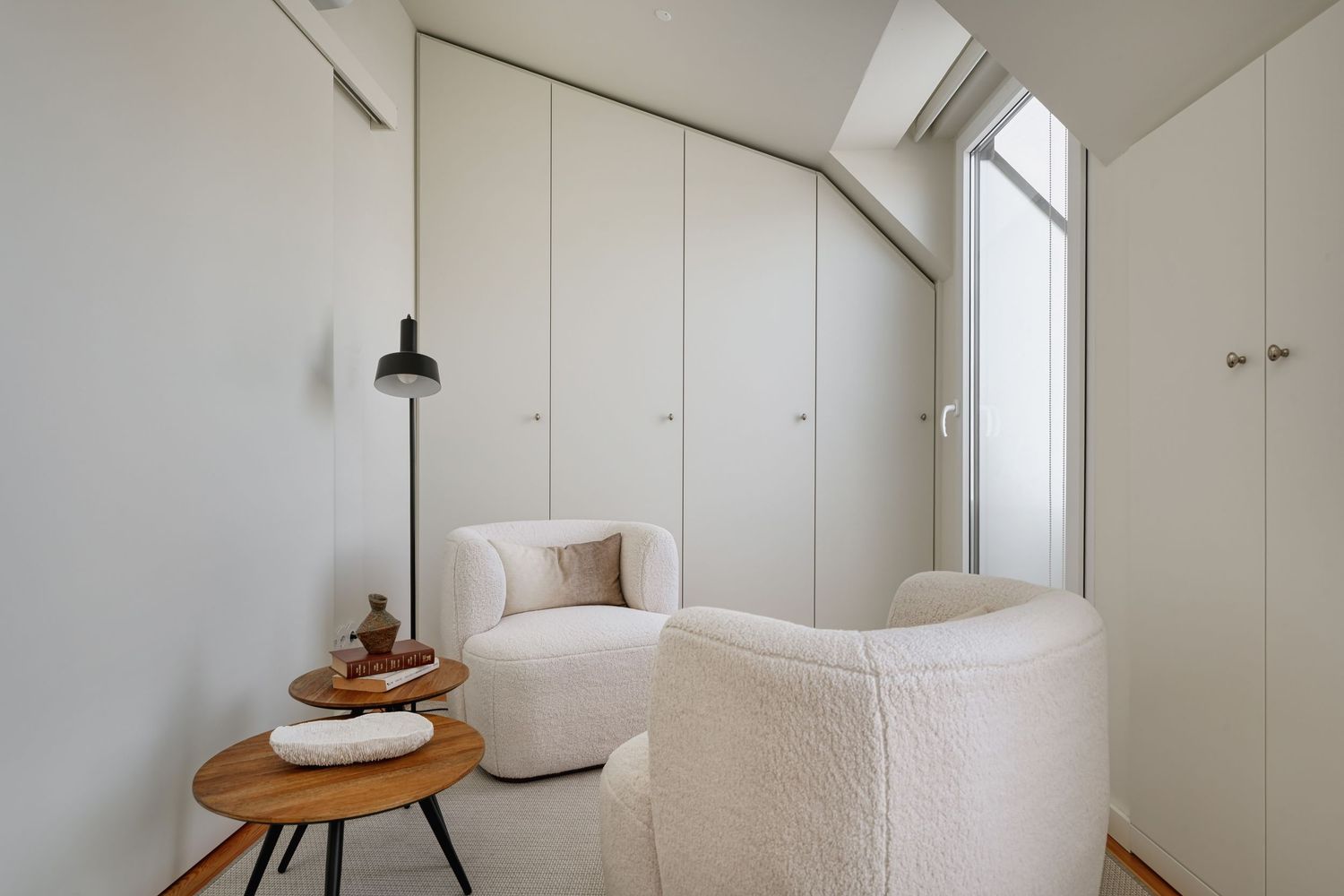 Minimalist white room with built-in wardrobe, two fluffy armchairs, wooden side table, and floor lamp under sloped ceiling.
