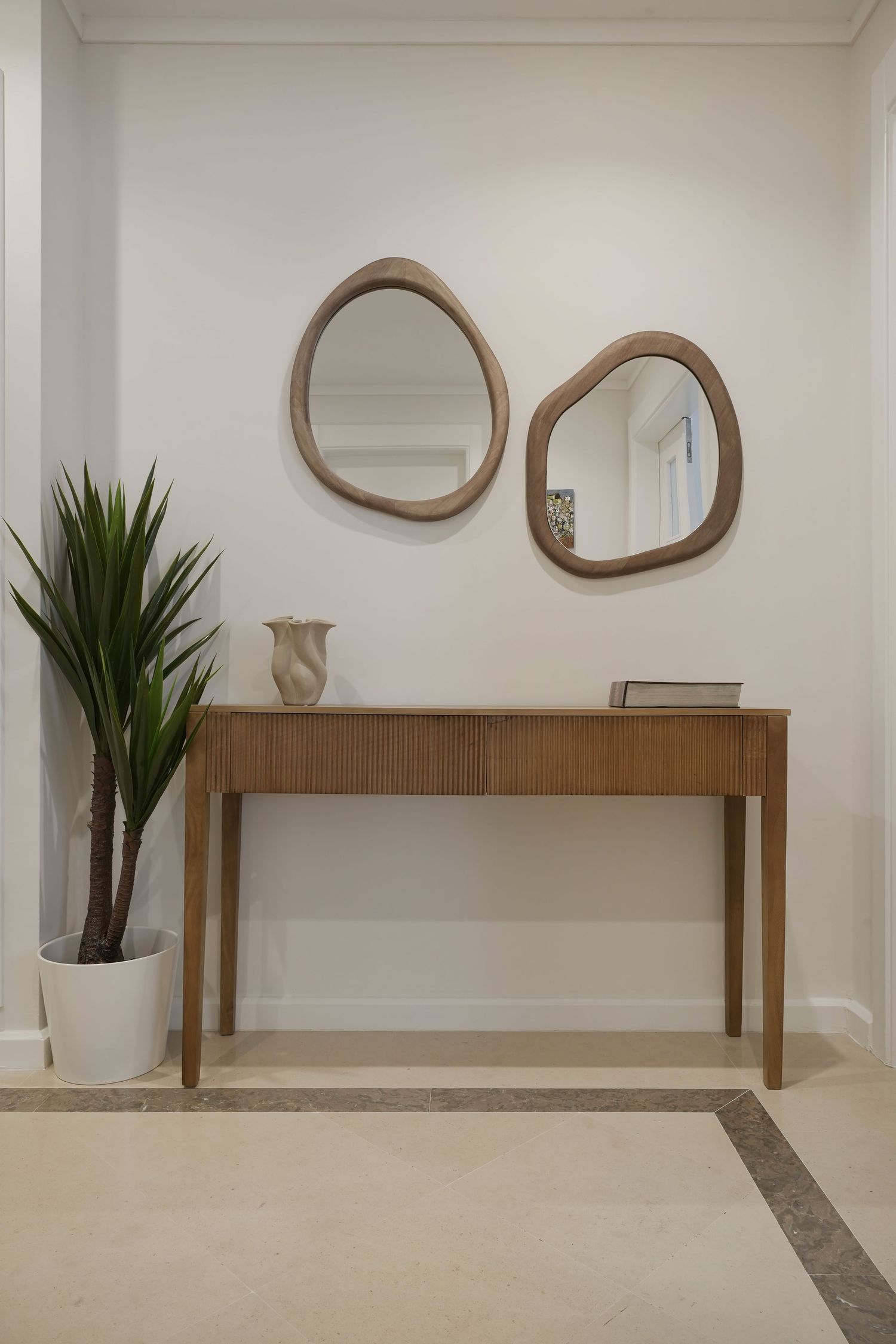Elegant entryway with wooden console table, two organic-shaped mirrors, ceramic vase, and potted plant on marble floor.
