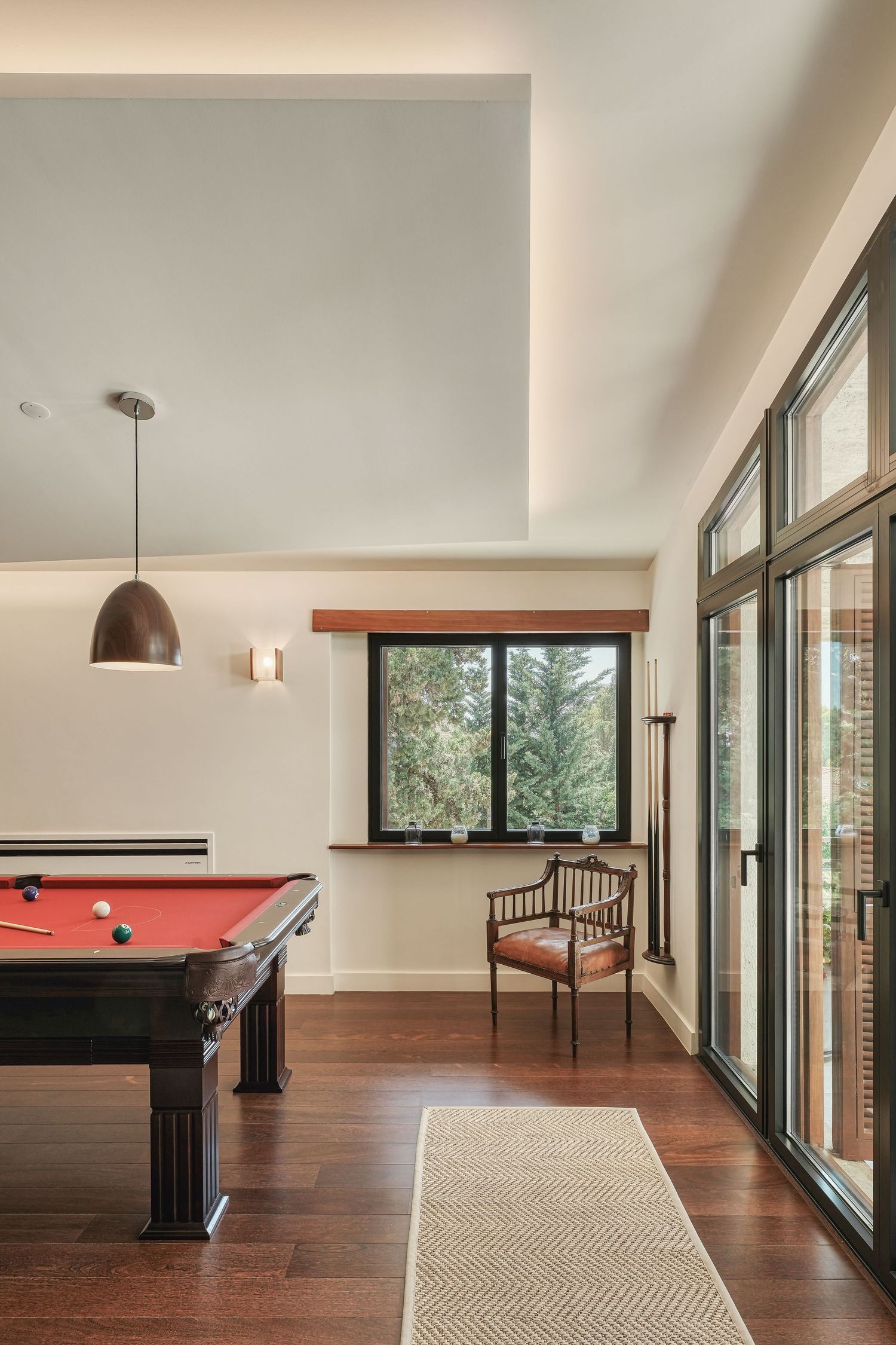 Modern game room with red felt pool table, hardwood floors, pendant light, and large windows overlooking trees.