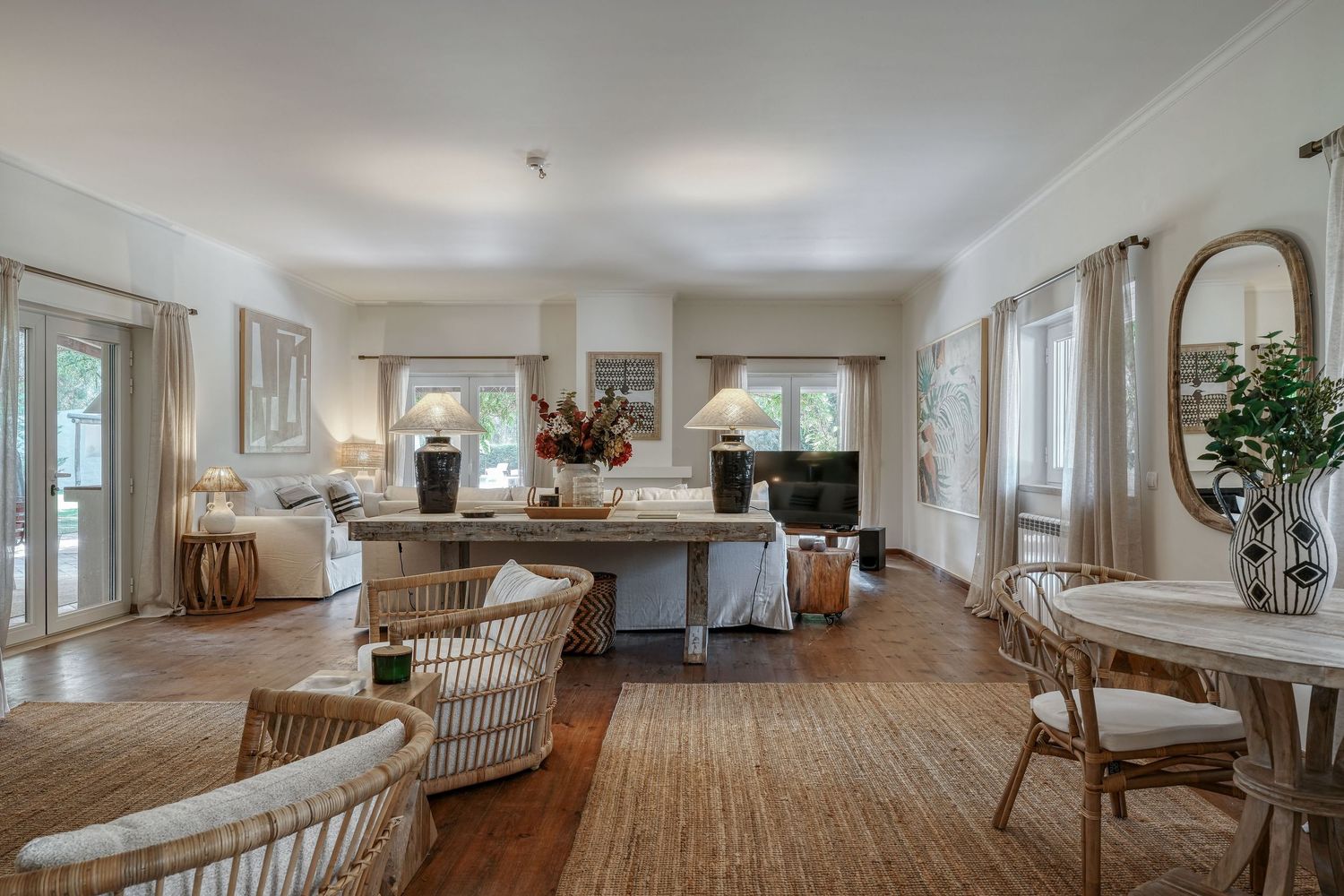 Bright open-plan living room with rattan furniture, hardwood floors, neutral tones, and large windows allowing natural light.