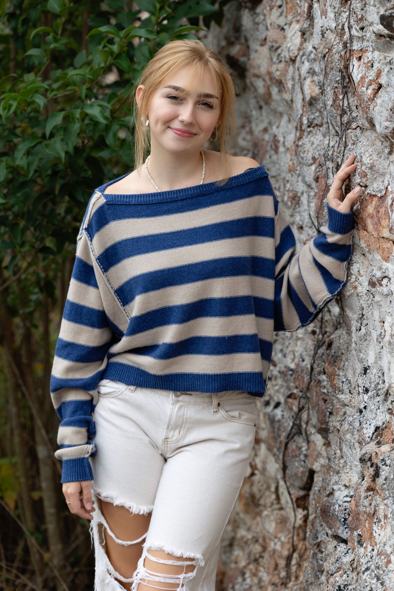 A person in a blue and beige striped sweater and white distressed jeans poses casually against a tree trunk outdoors.