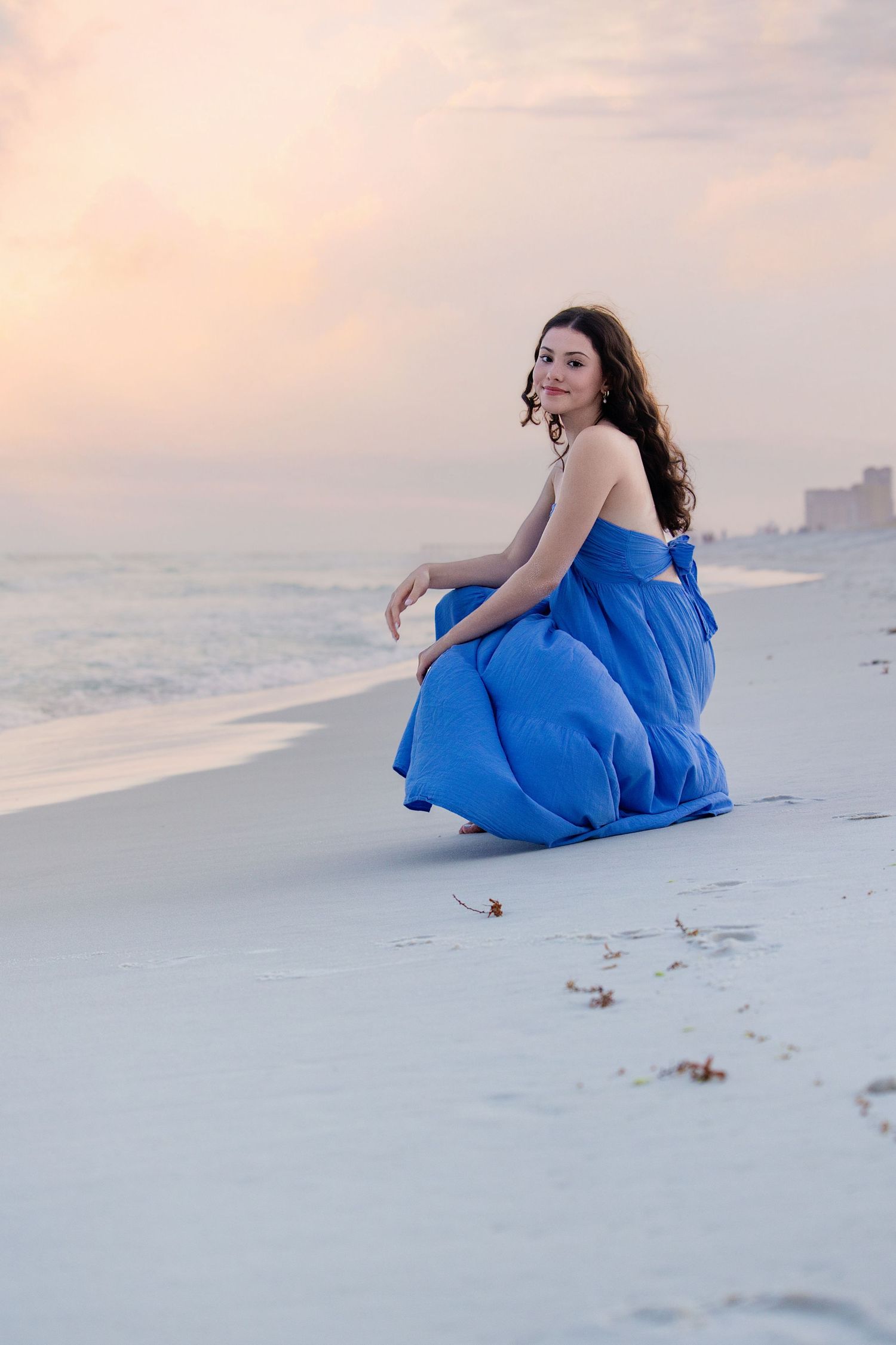 Figure in flowing blue dress reclining on white sandy beach at sunset.