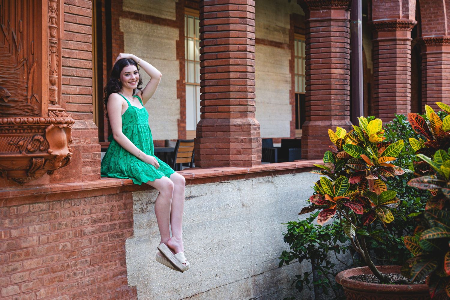 A person in a green sundress sits on a brick ledge near colorful potted plants outside a historic building.