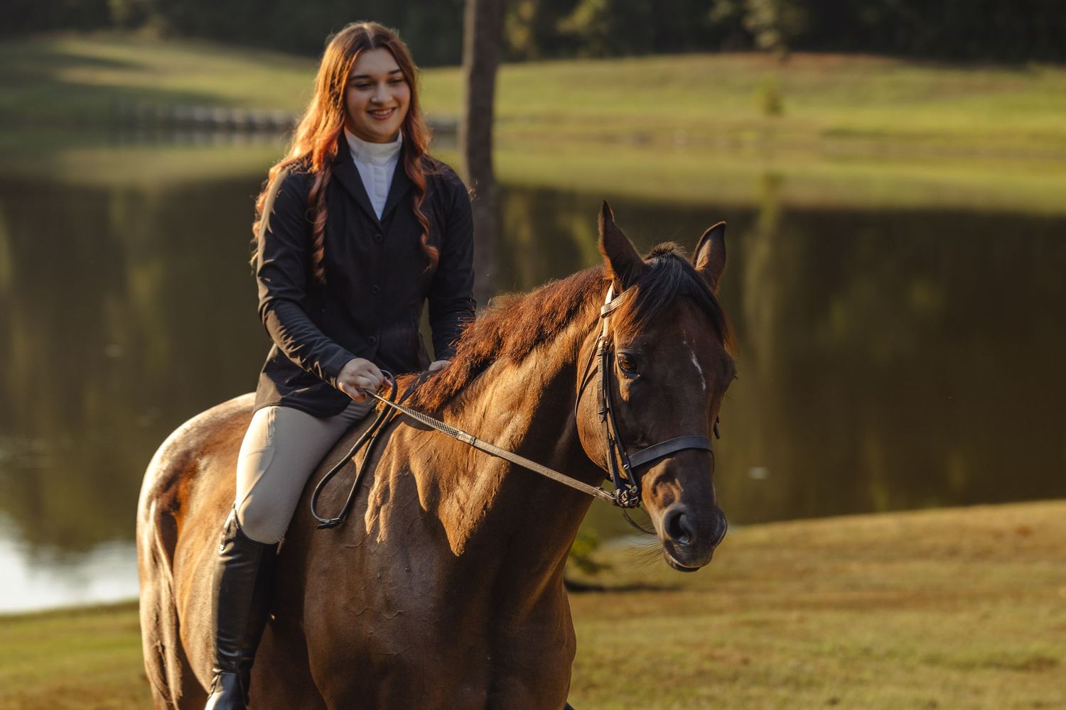 Young woman with red hair in equestrian attire riding a chestnut horse near a pond in golden hour light.