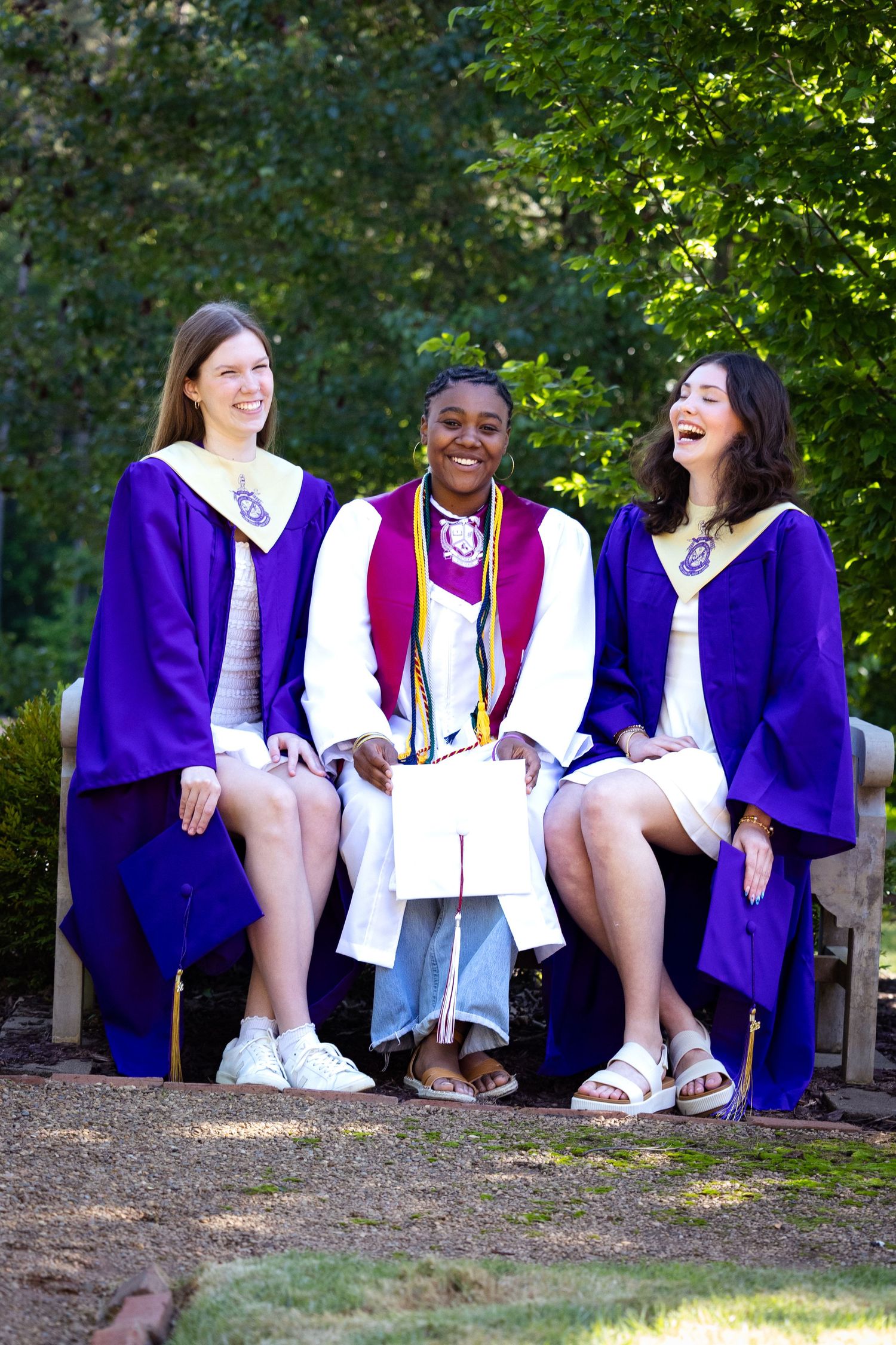 Three smiling graduates in purple robes sit on a bench outdoors celebrating their graduation day.