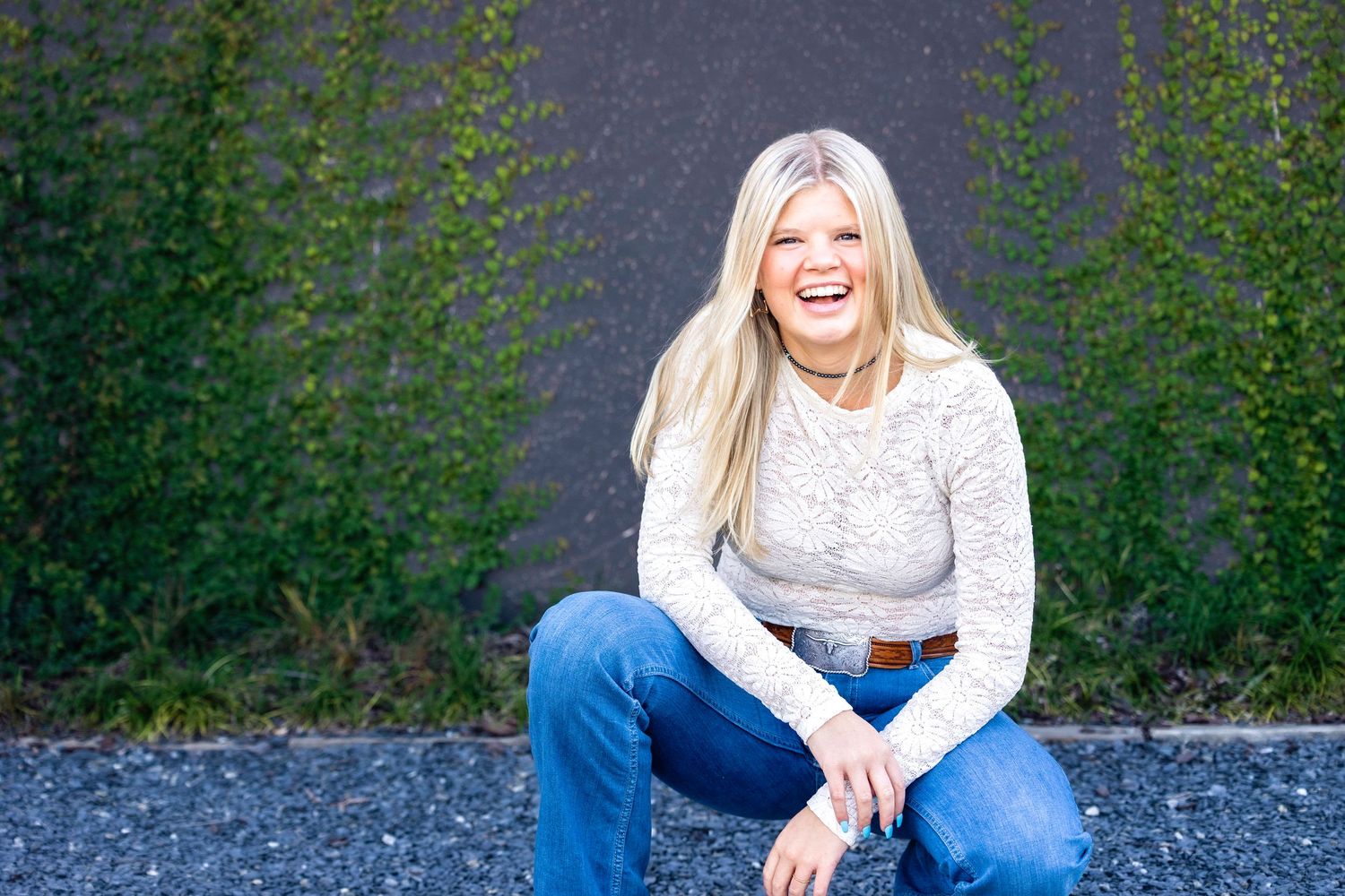 A person in blue jeans and white sweater sits laughing against a dark wall with green vines growing behind them.