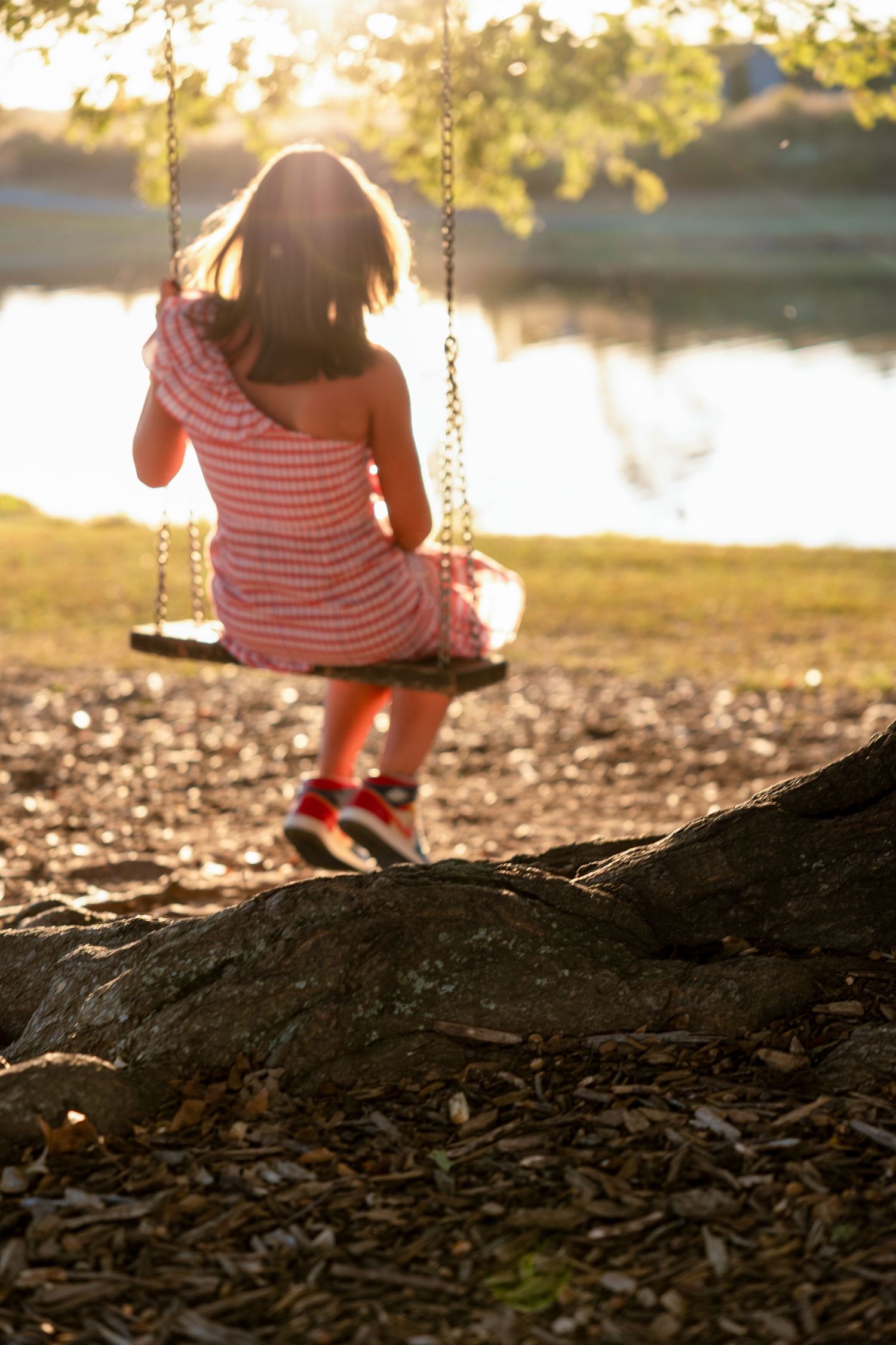 A person in a striped outfit sits on a wooden swing at sunset by a lake with golden light filtering through trees.