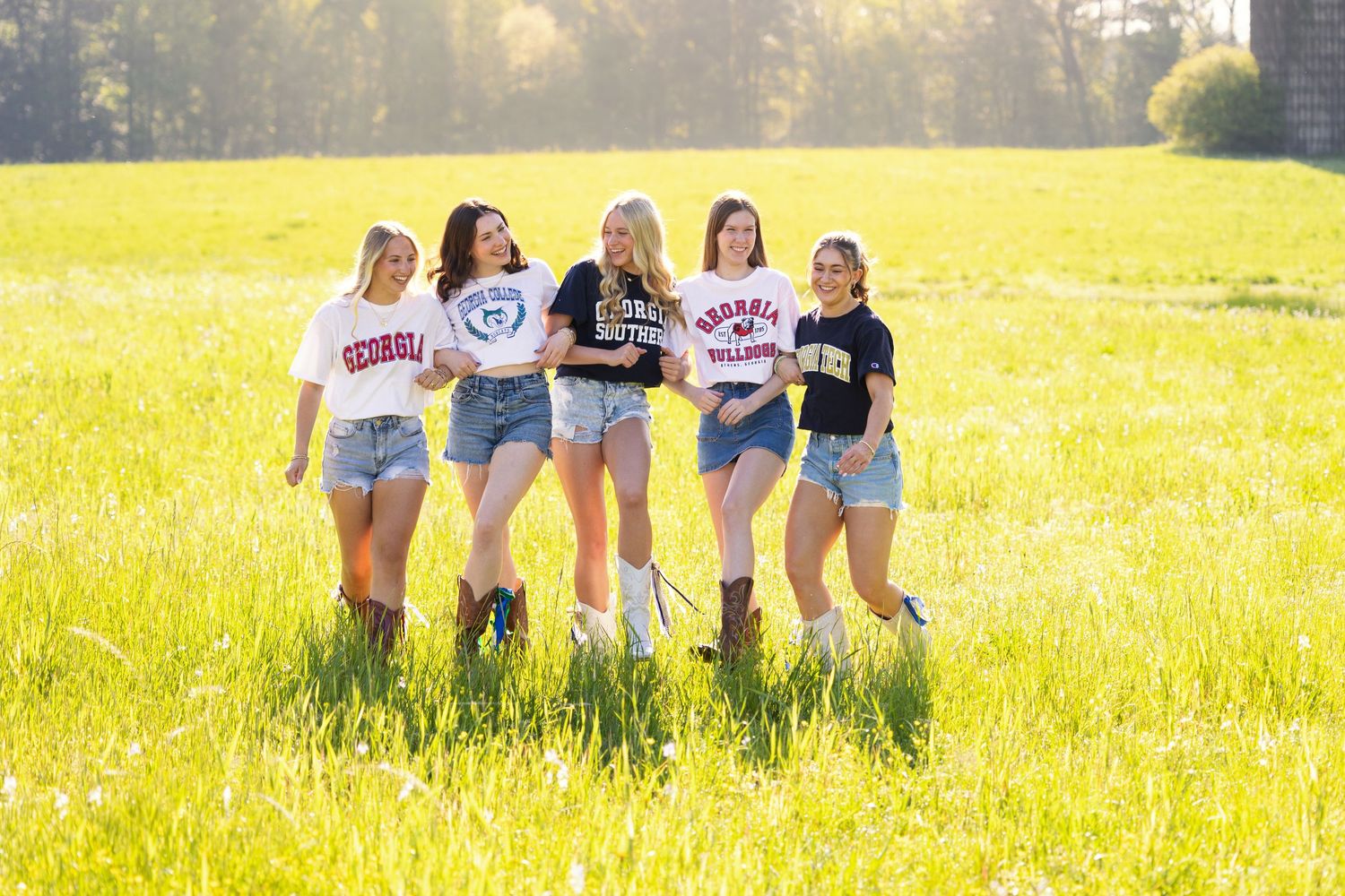 Five young women in college shirts and denim shorts pose in a sunny green field wearing cowboy boots.
