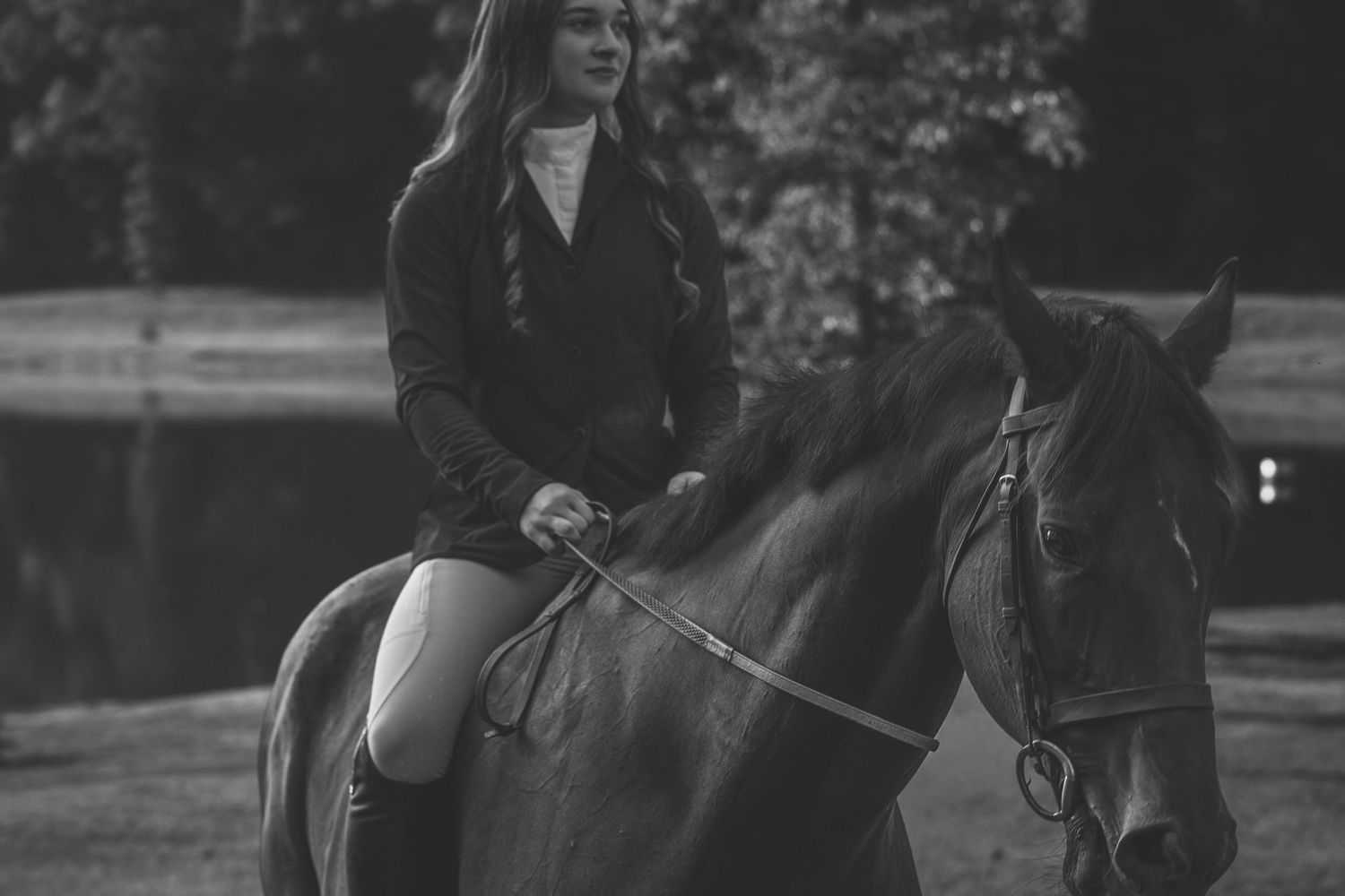 Black and white photo of a young woman in equestrian attire riding a dark horse outdoors.