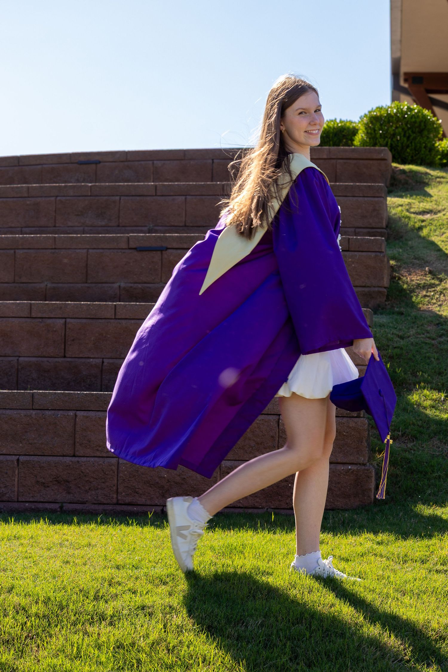Young woman walking on grass in purple graduation gown and cap, smiling outdoors near brick wall on sunny day.