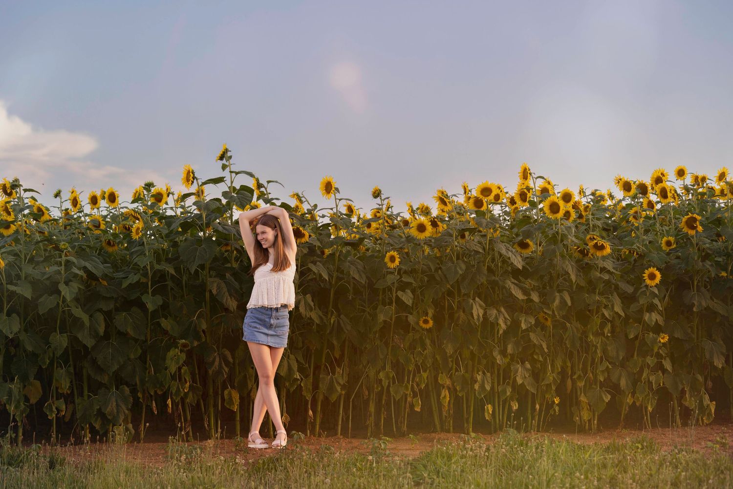 A person stands in front of a tall sunflower field at sunset with arms raised in a carefree pose.