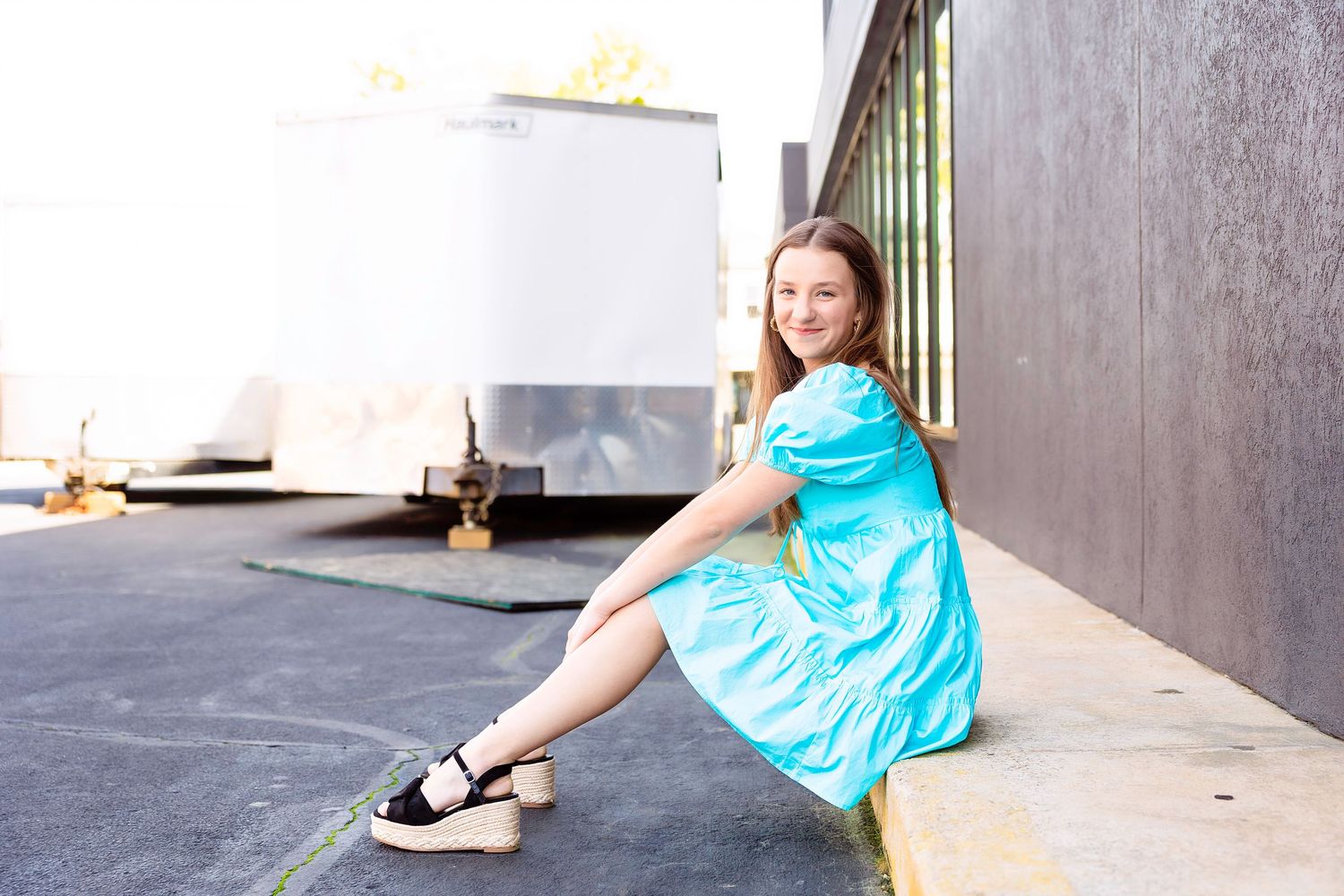 Someone in a turquoise summer dress and black sandals sits casually on a concrete ledge against an industrial backdrop.