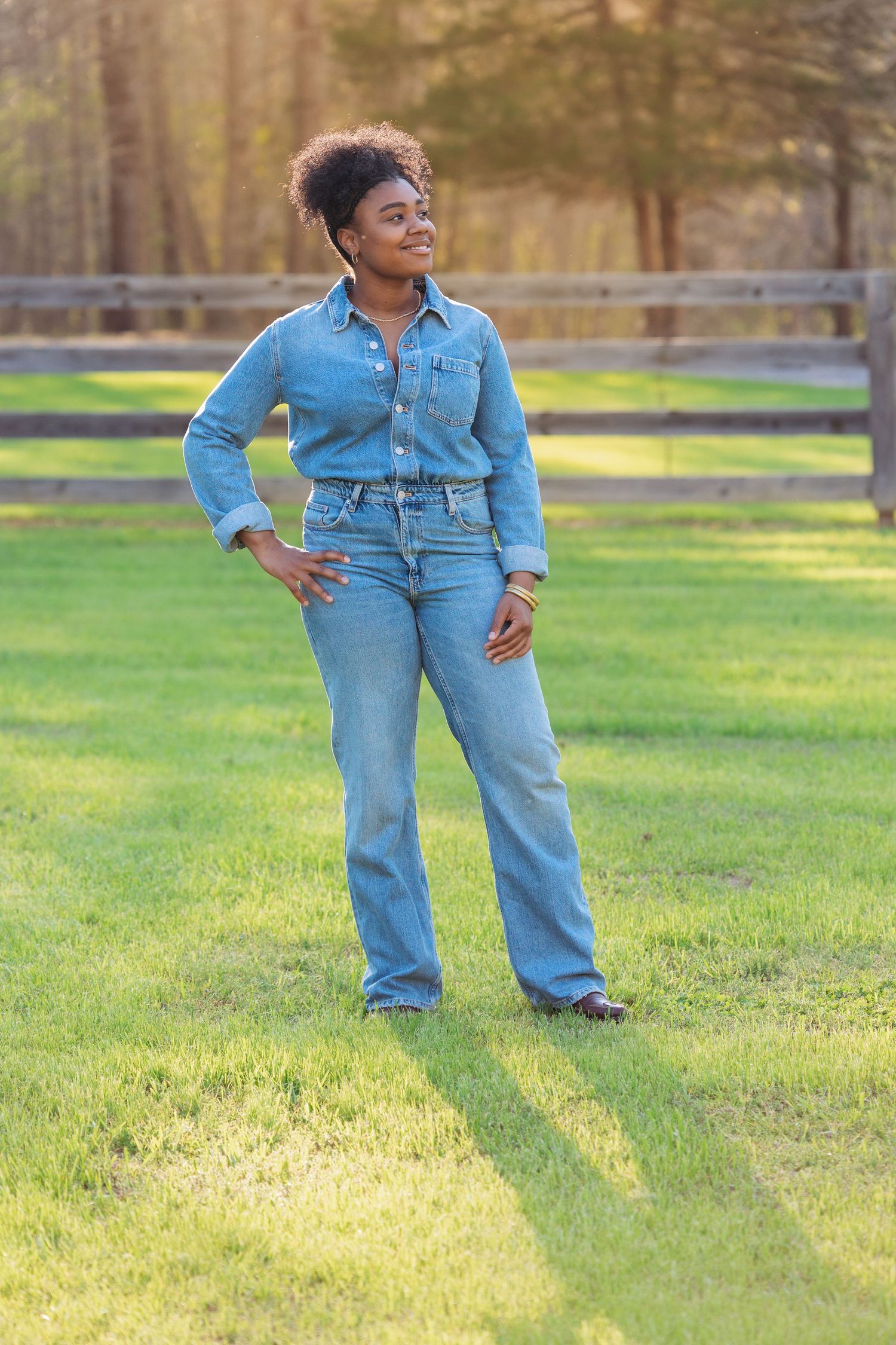 Woman in denim shirt and jeans standing on green grass near wooden fence in golden hour sunlight.