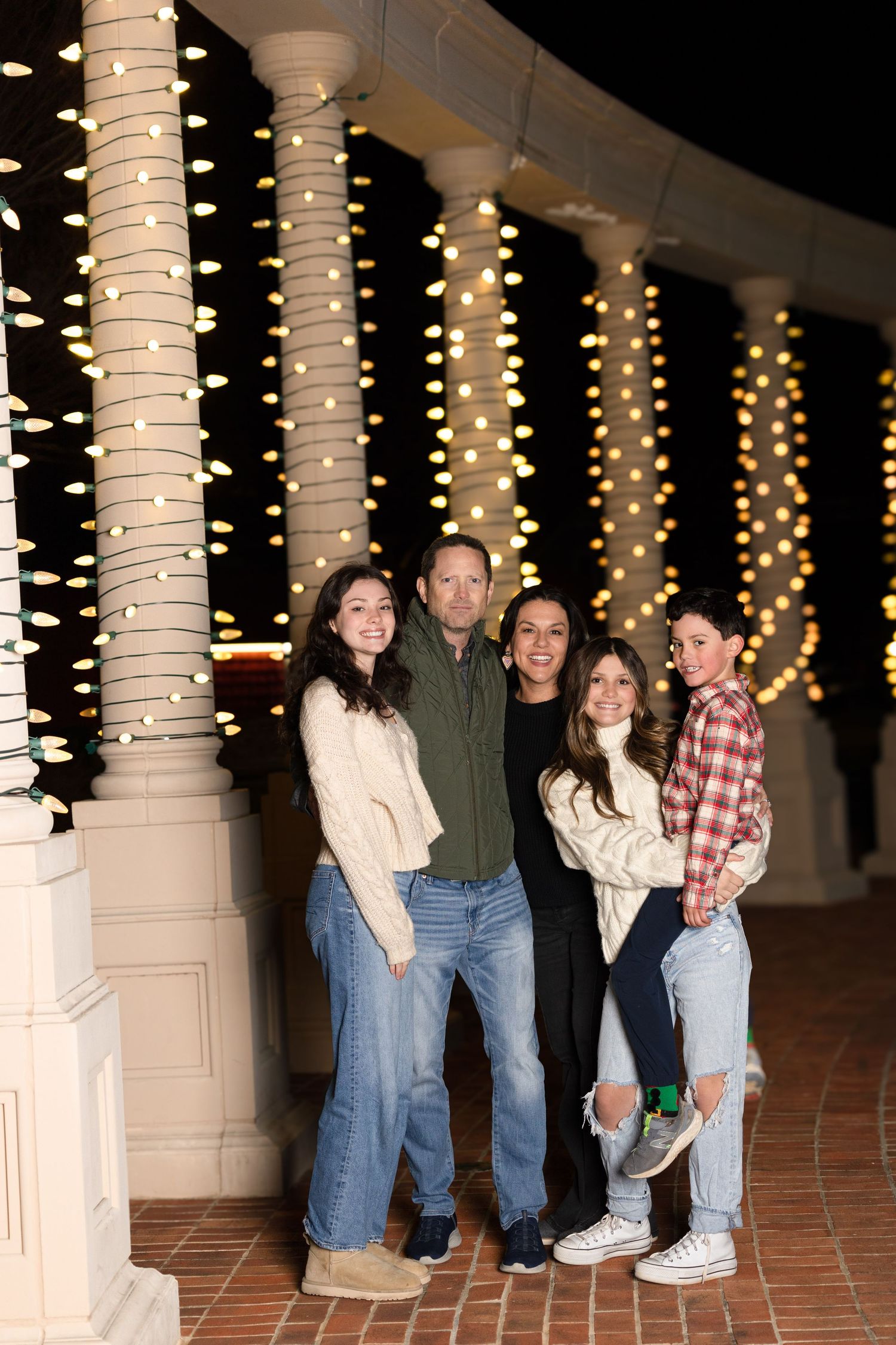 Family poses together against white columns decorated with twinkling Christmas lights at night.