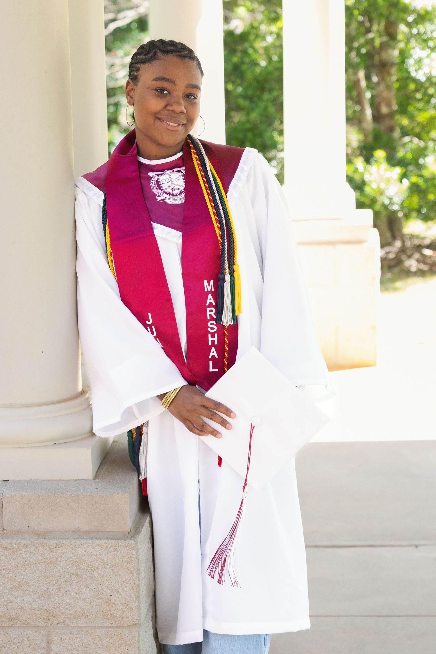 Young woman in white graduation gown with marshal sash smiling outdoors near columns.