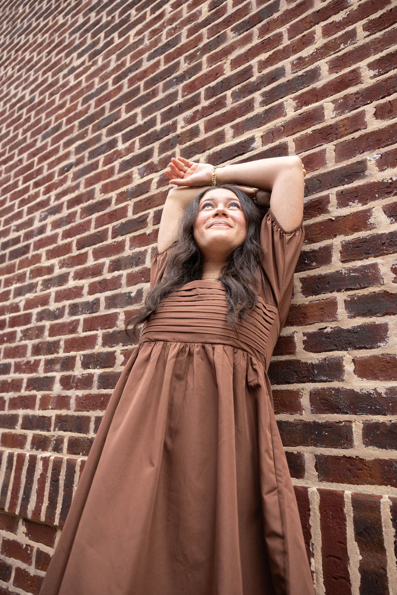 A woman in a brown dress poses expressively against a rustic brick wall in a photo series.
