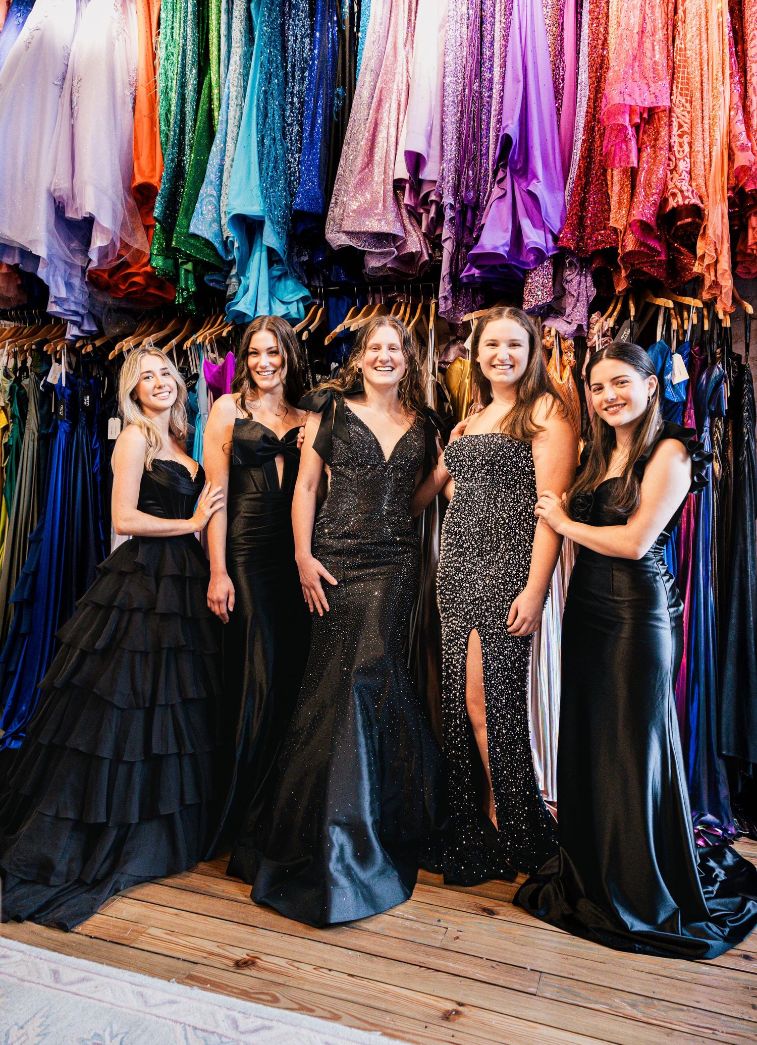 A row of formal dresses in black and sparkly patterns hangs on display against a colorful backdrop of evening gowns.