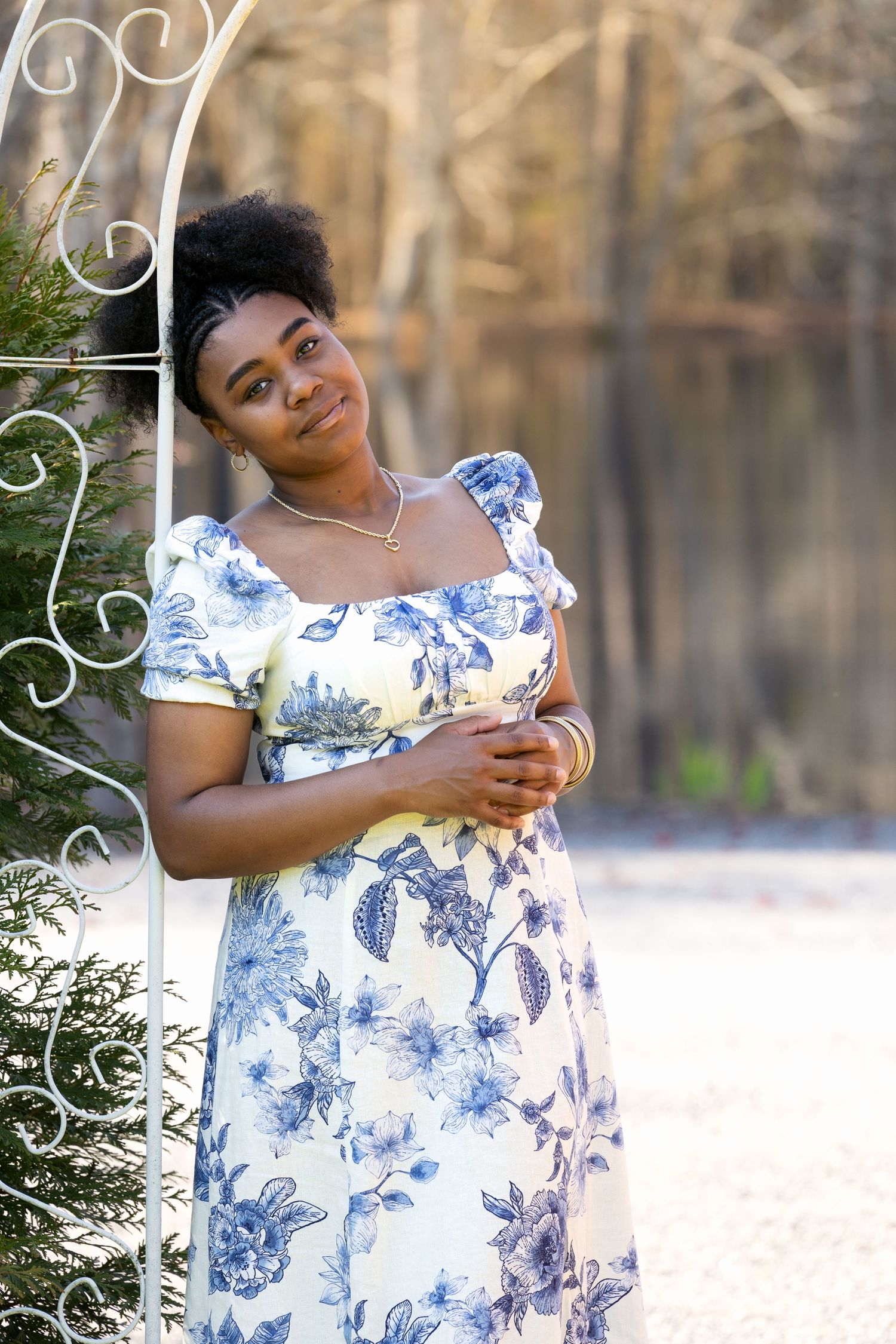 Young Black woman in blue floral dress leaning against white iron arbor in outdoor garden setting.