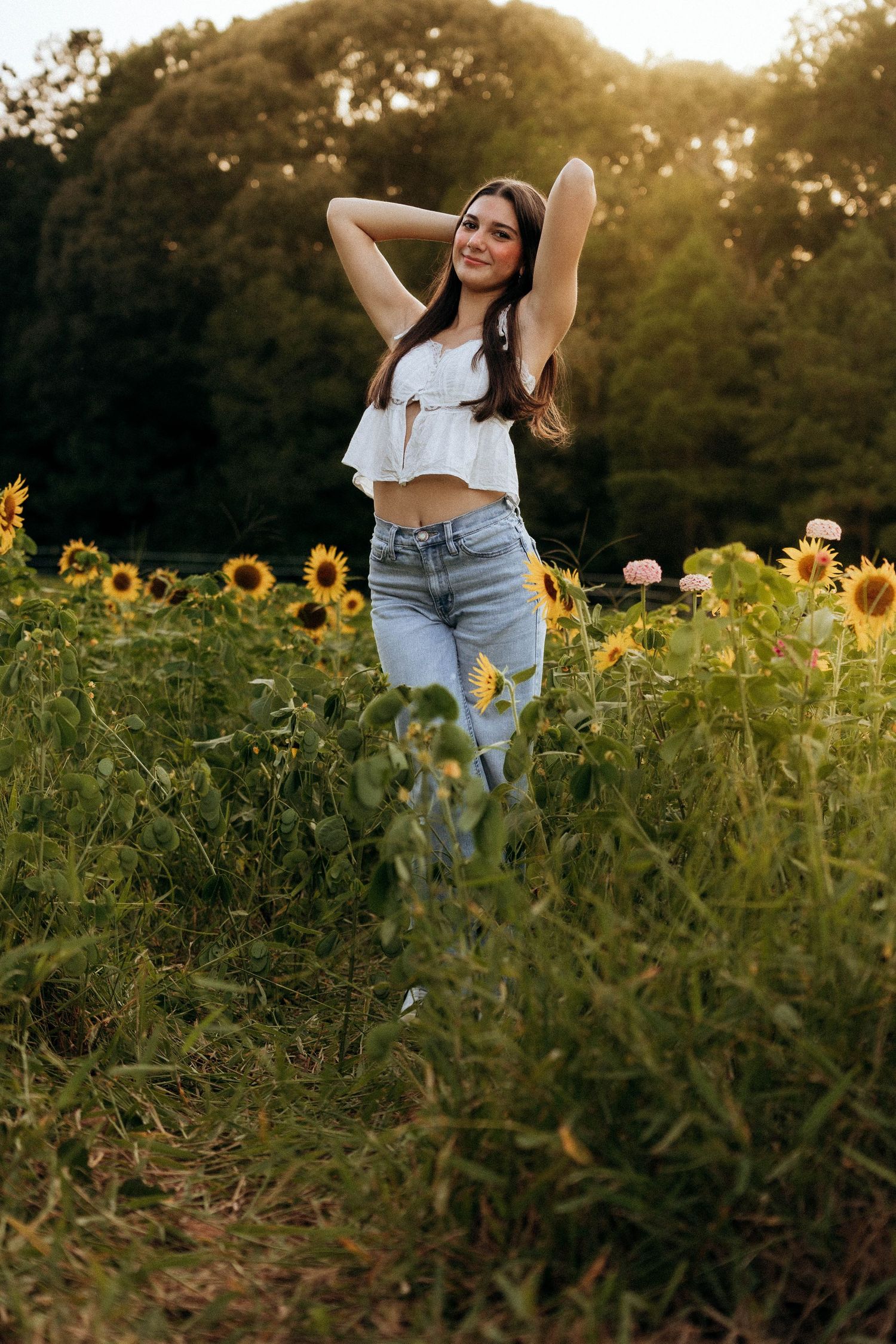 A figure stands among blooming sunflowers in a field at golden hour wearing denim jeans and a white top.