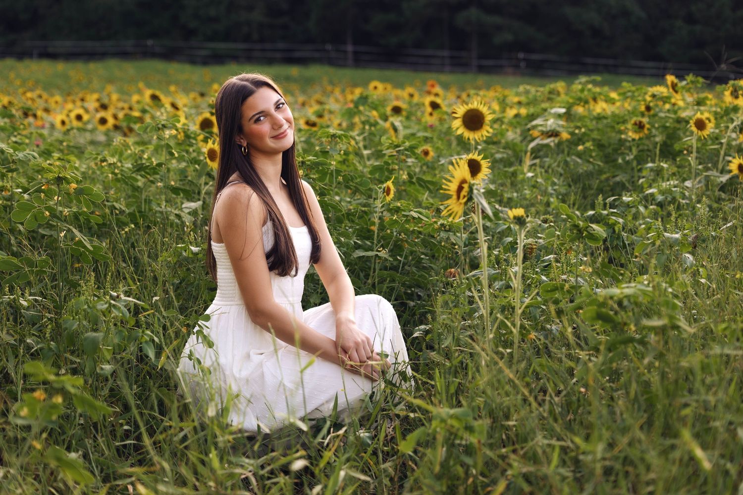 Woman in white dress sitting among sunflowers during sunset.