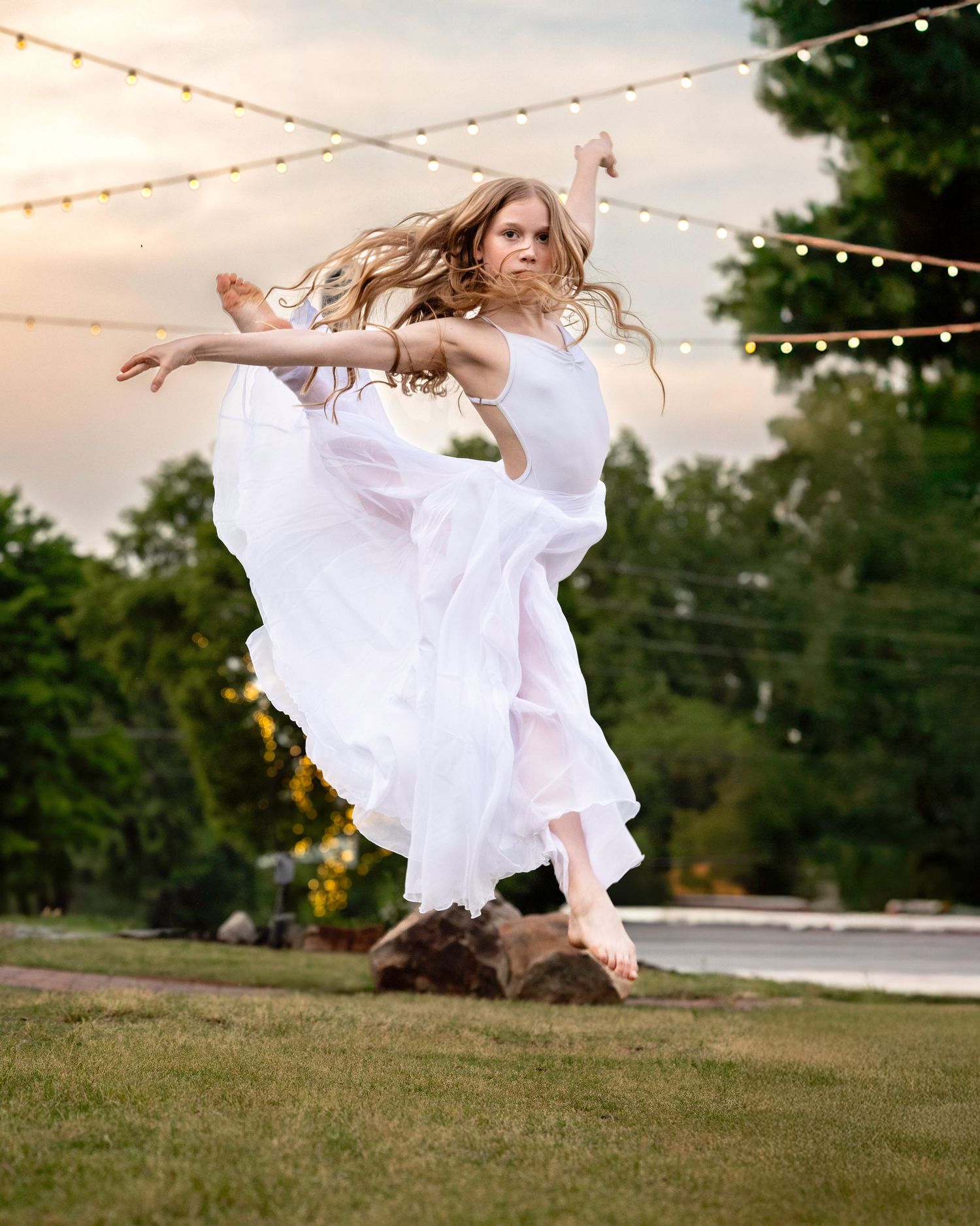 Figure in white flowing dress spins and twirls under twinkling string lights at dusk.