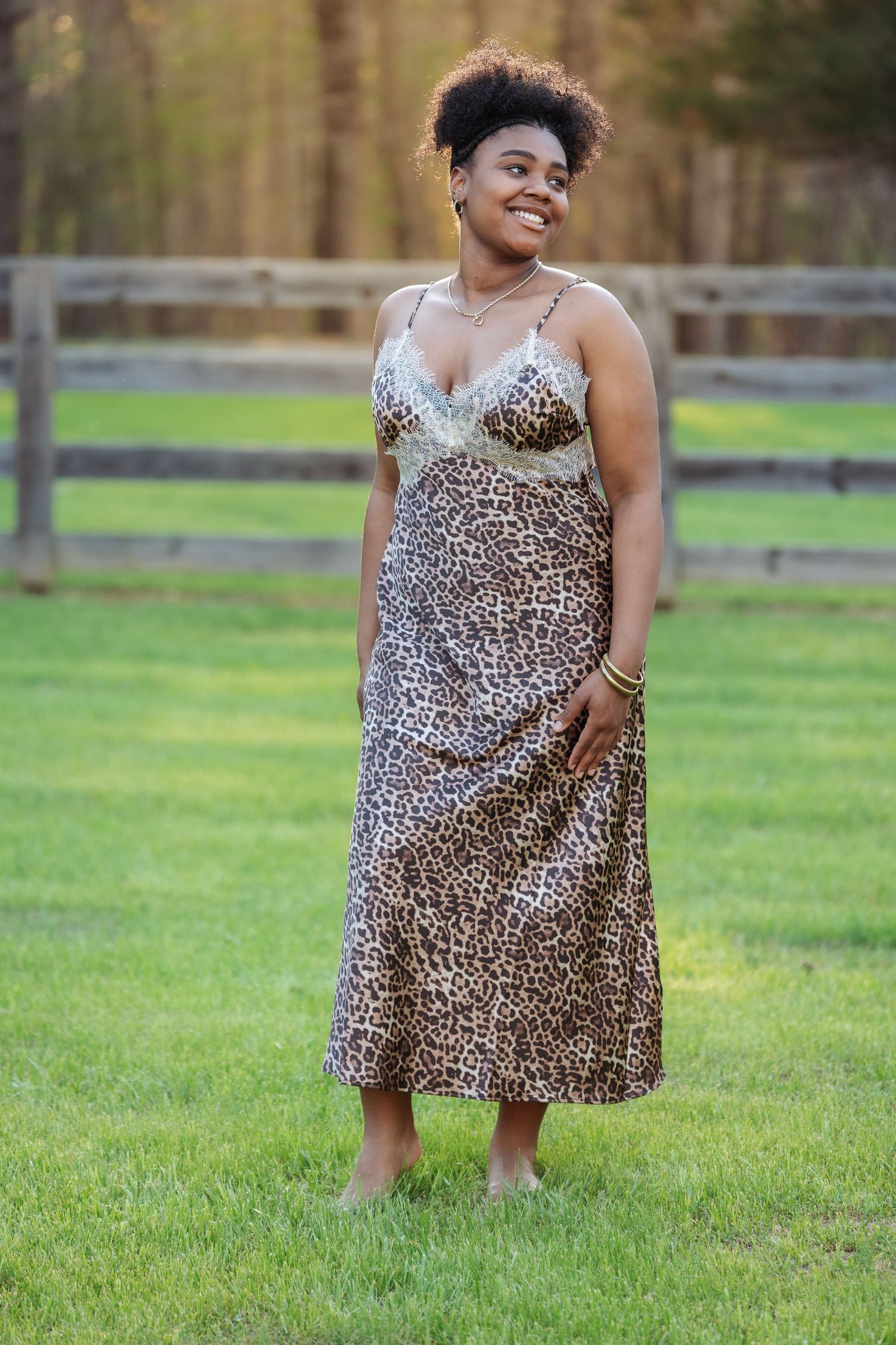 Woman in leopard print midi dress standing on green grass outdoors near wooden fence, smiling confidently.