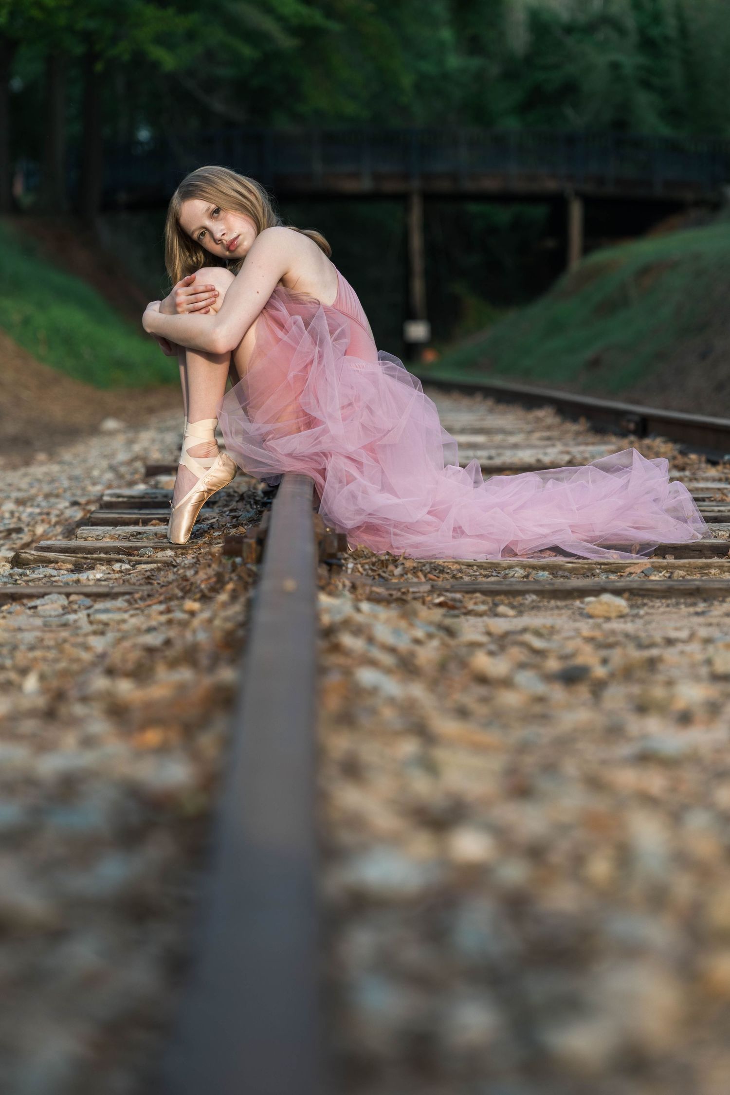 A figure in a flowing pink tulle dress poses gracefully along train tracks during an outdoor photo session.