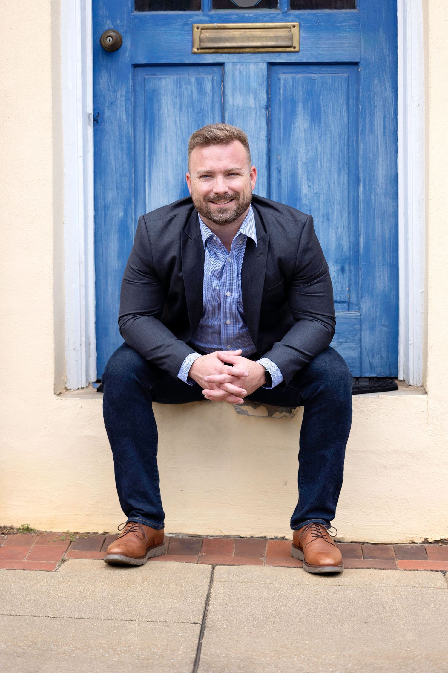 Professional in a dark suit sits casually against blue doors of a historic building in an outdoor portrait session.
