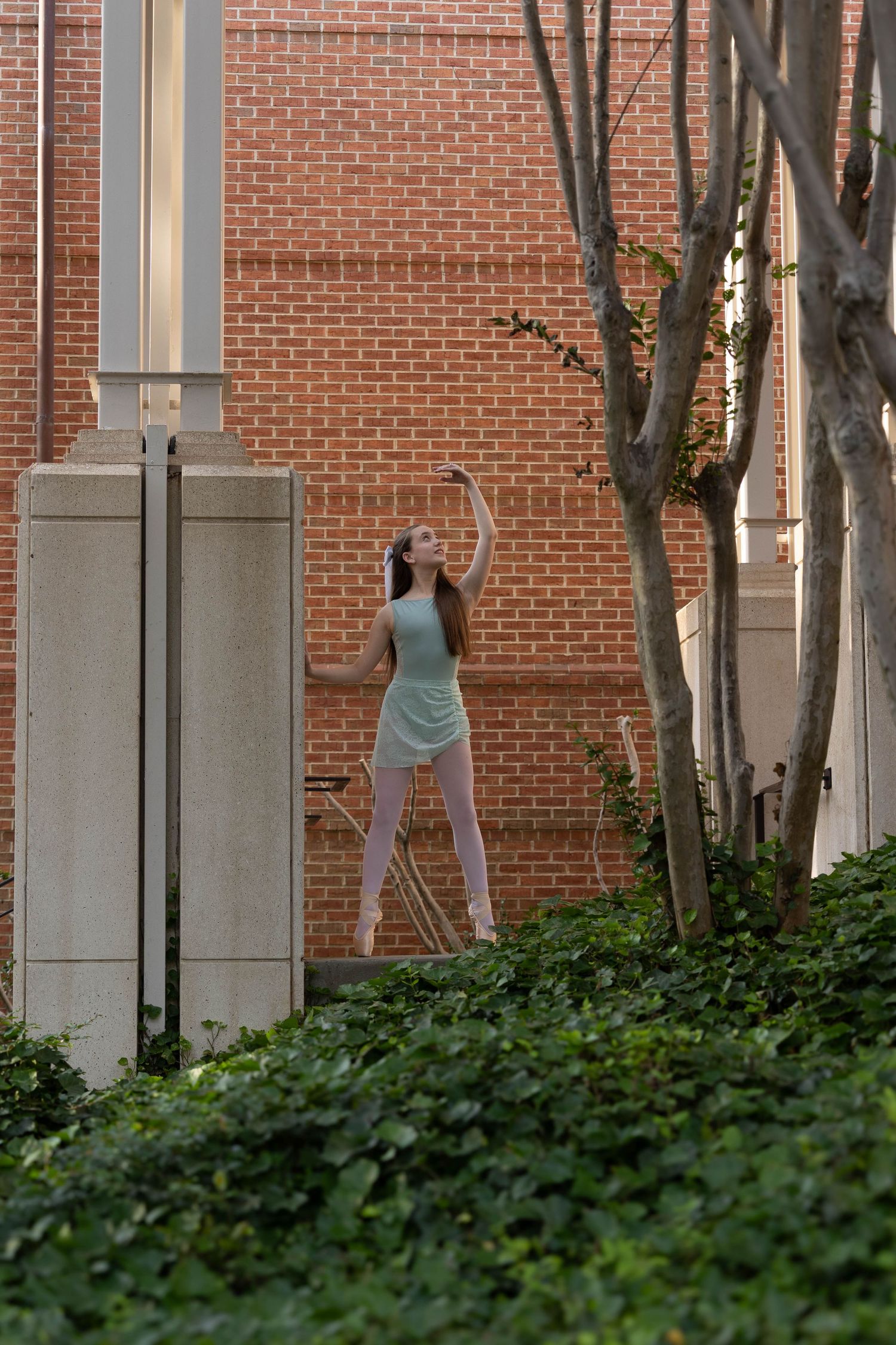 A person in a casual dress poses against a brick wall near ivy-covered ground and trees at sunset.