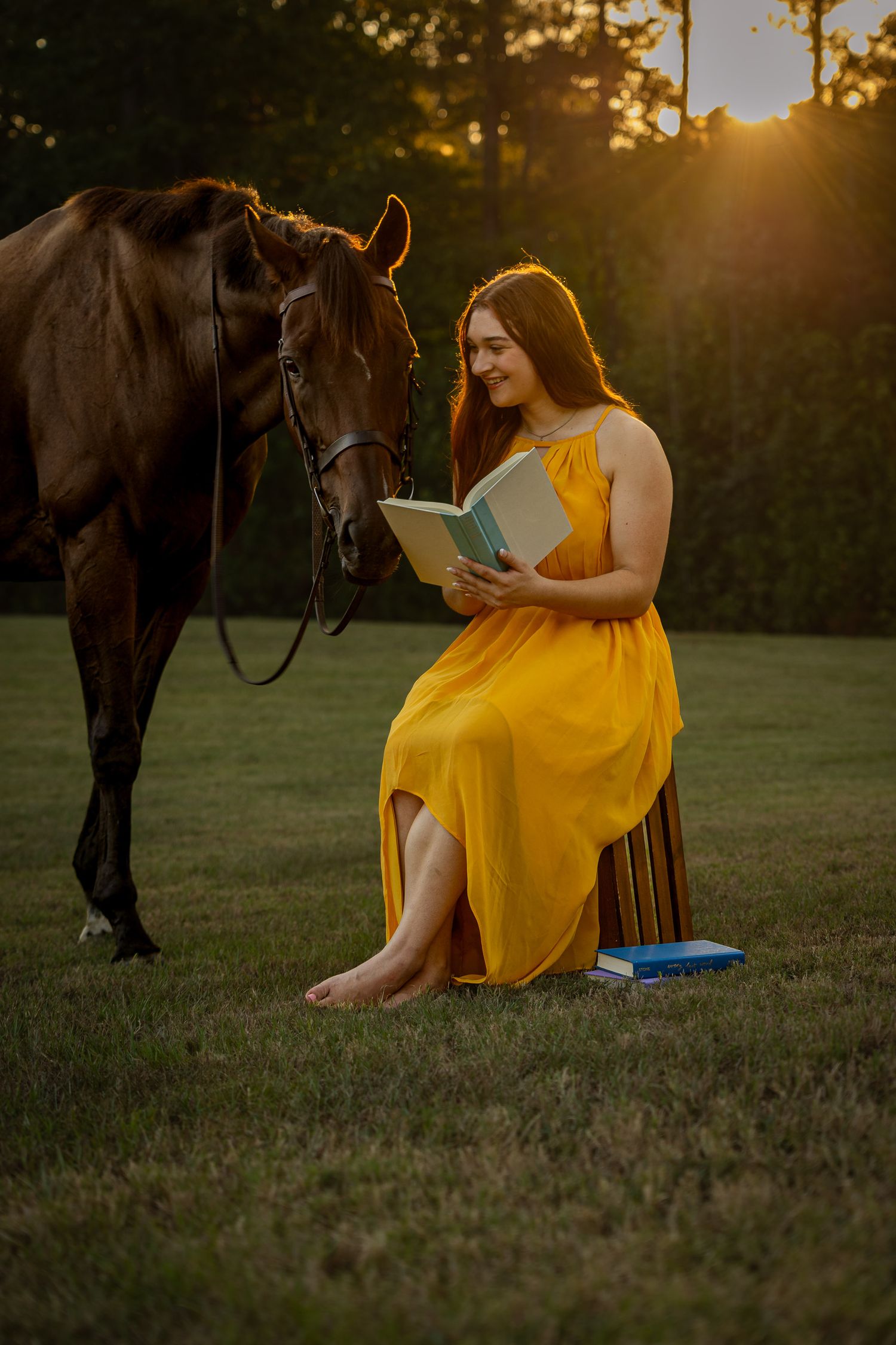 Woman in yellow dress reads book beside brown horse at golden sunset outdoors.