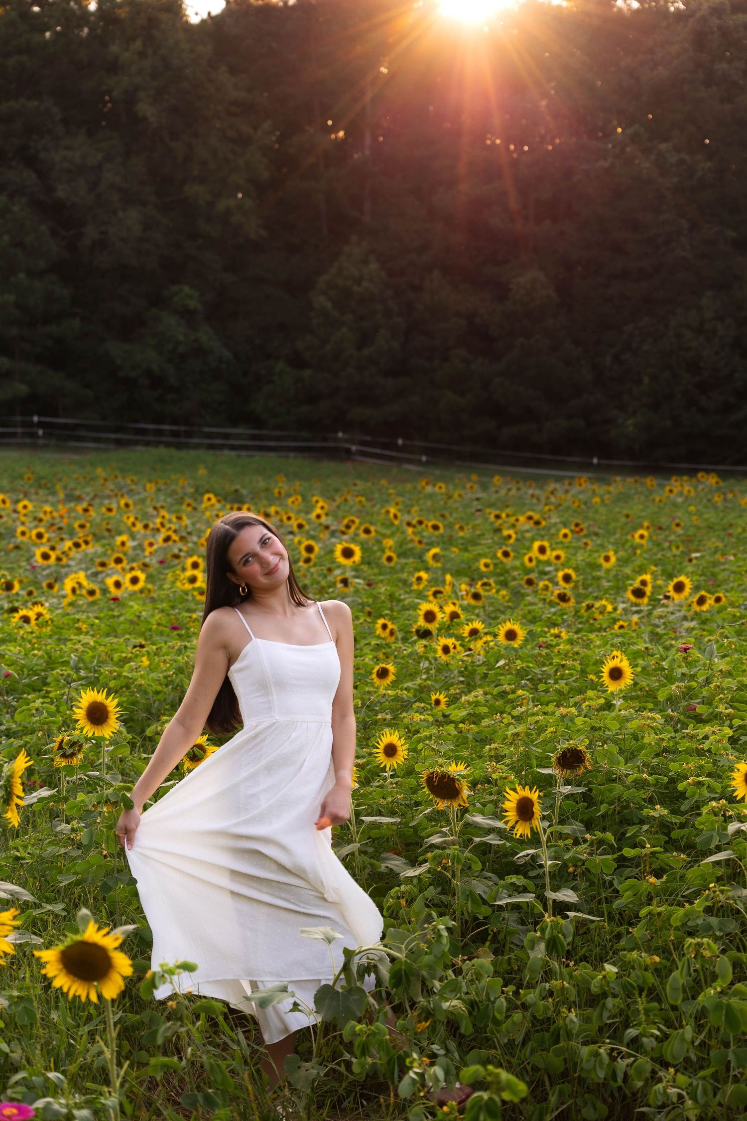 Woman in flowing white dress twirling in sunflower field at sunset.