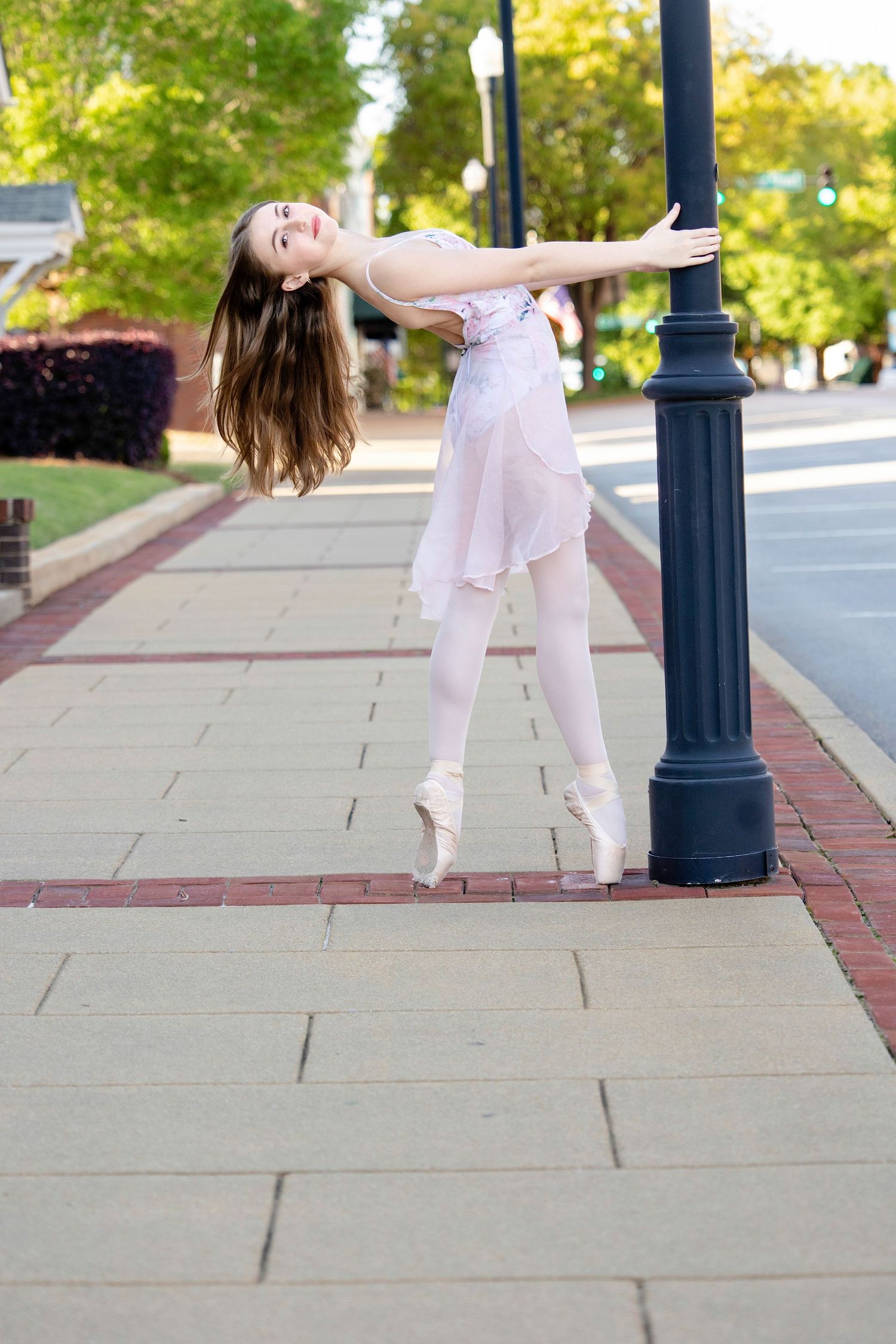 Dancers in white performing graceful movements along a sidewalk with decorative lampposts.