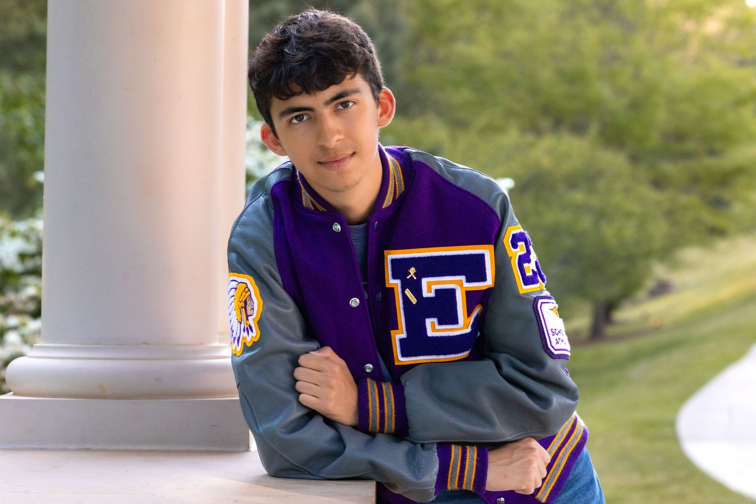 Teen boy in purple and gray varsity letterman jacket leaning against white column outdoors.