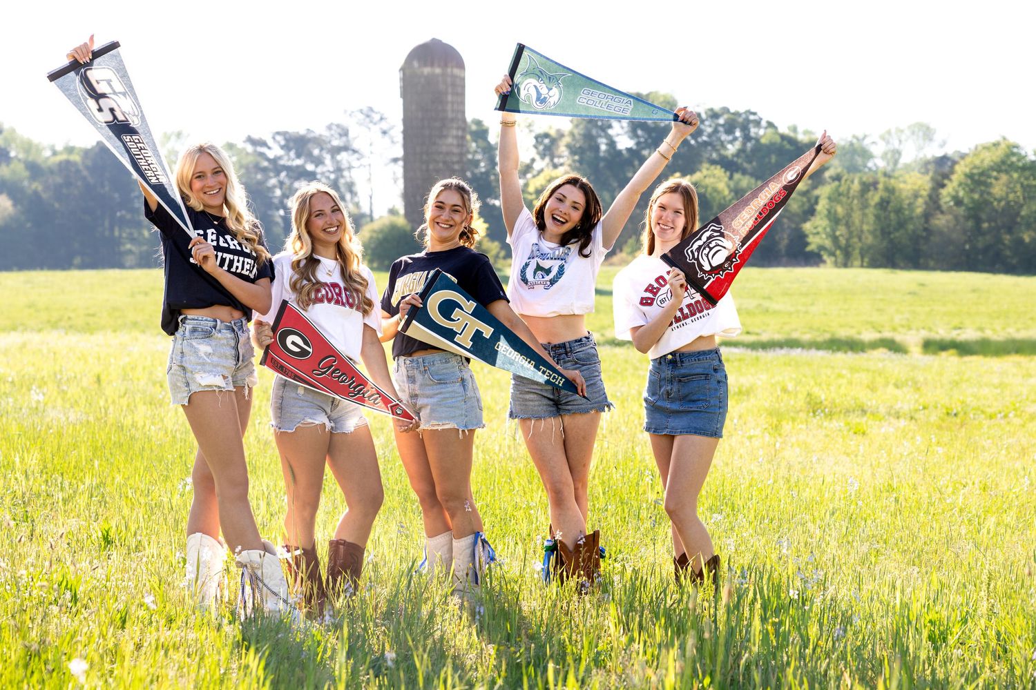 Five young women celebrate college commitments holding pennants and flags in a sunny green field.