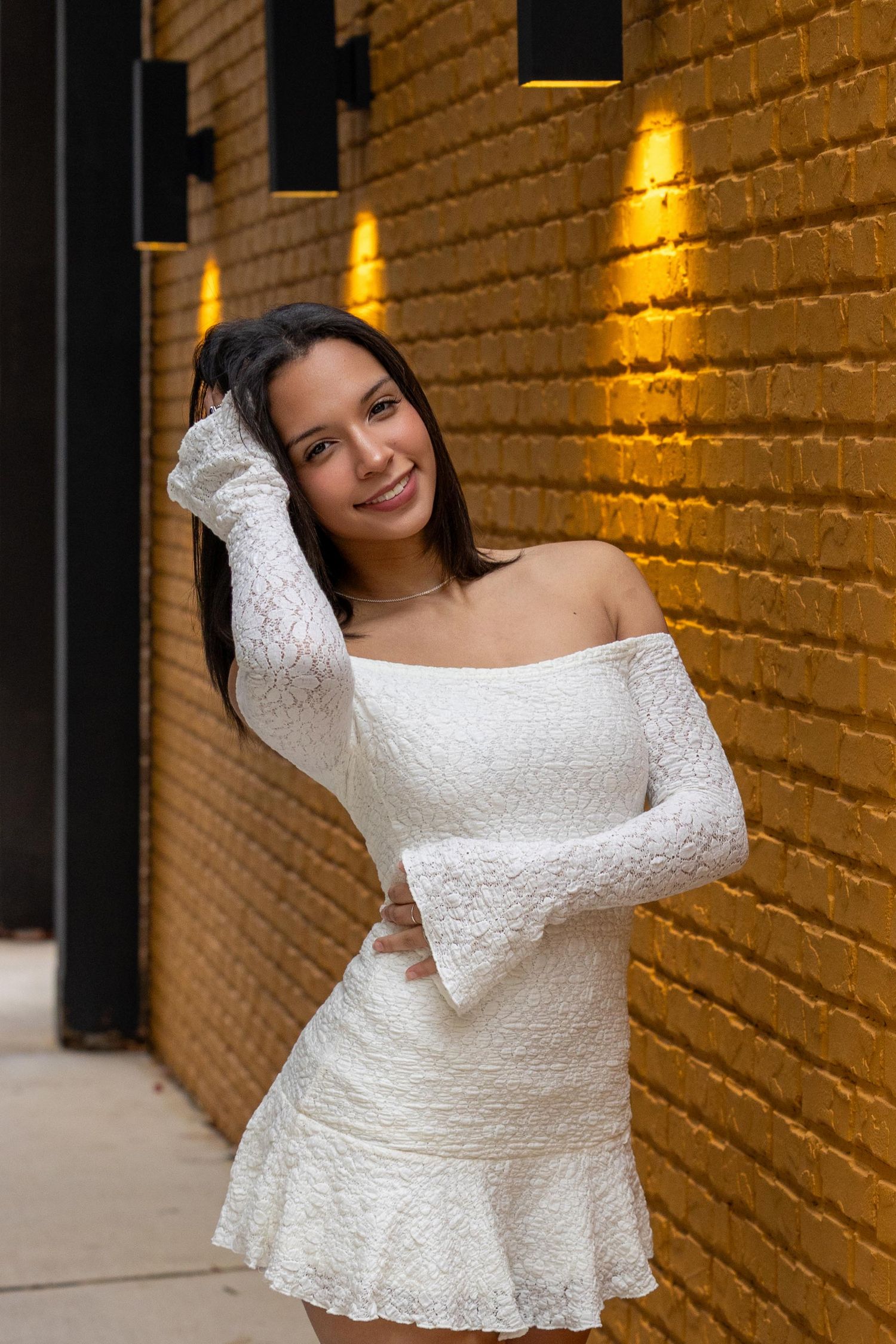 Person wearing an elegant white lace off-shoulder top poses against a brick wall with warm lighting.