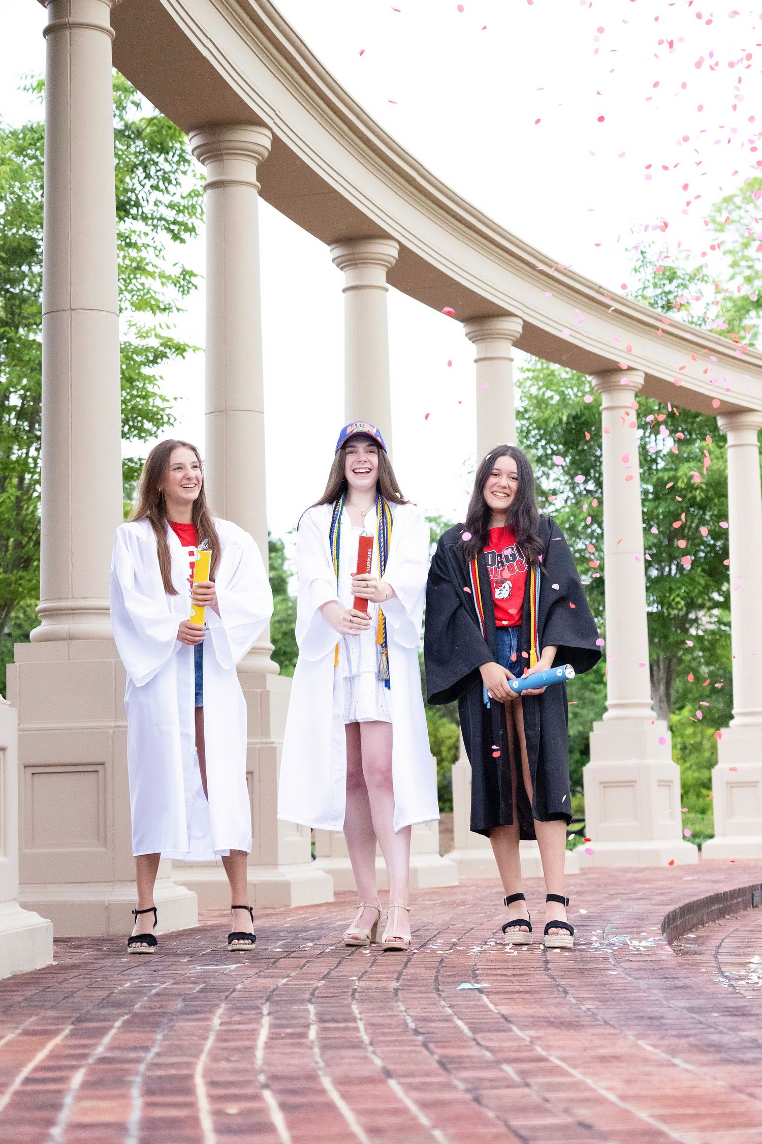 A group of graduates in white dresses and black gowns pose together under a curved colonnade walkway.