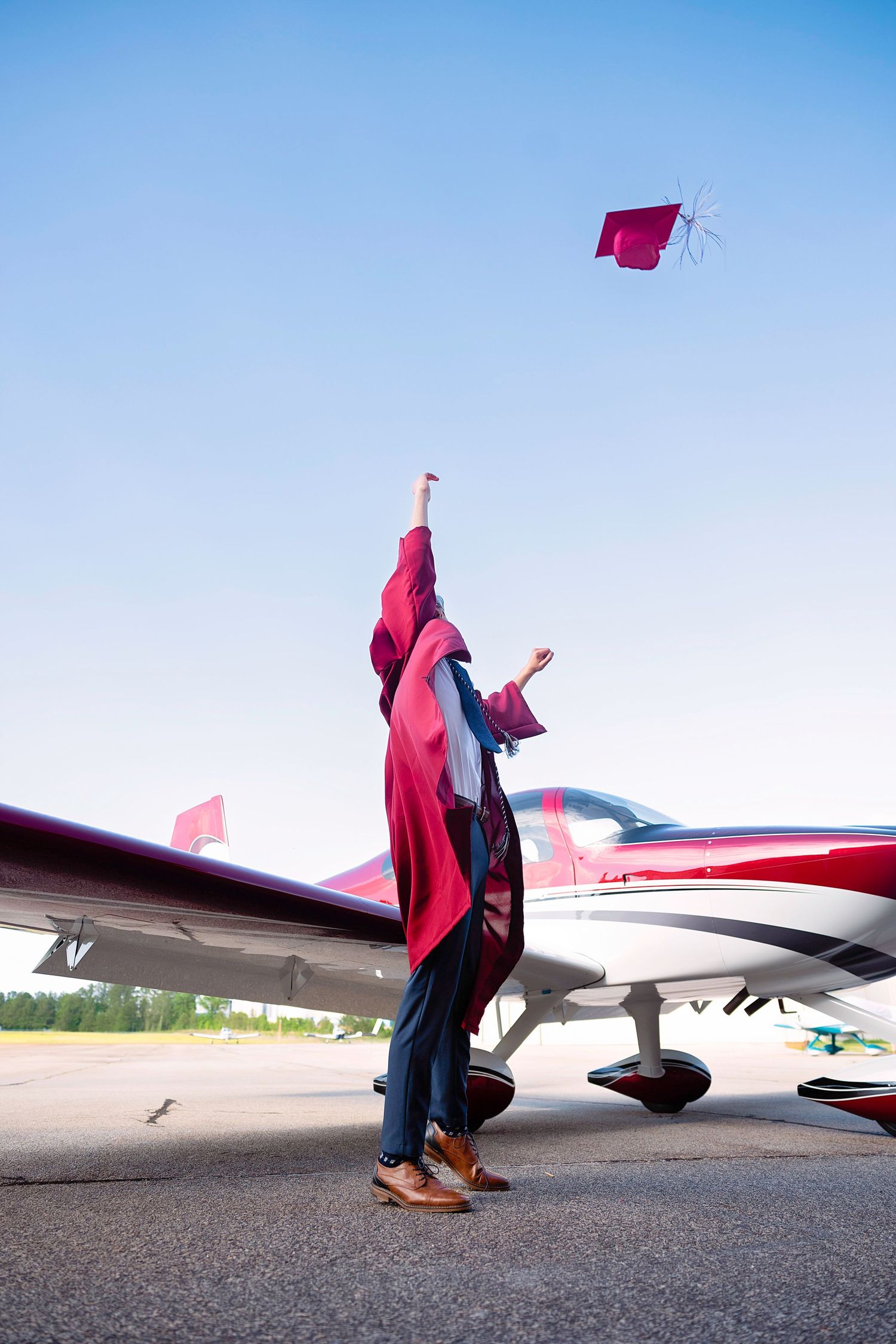 Graduation gown flowing dramatically beside a parked aircraft on a runway.
