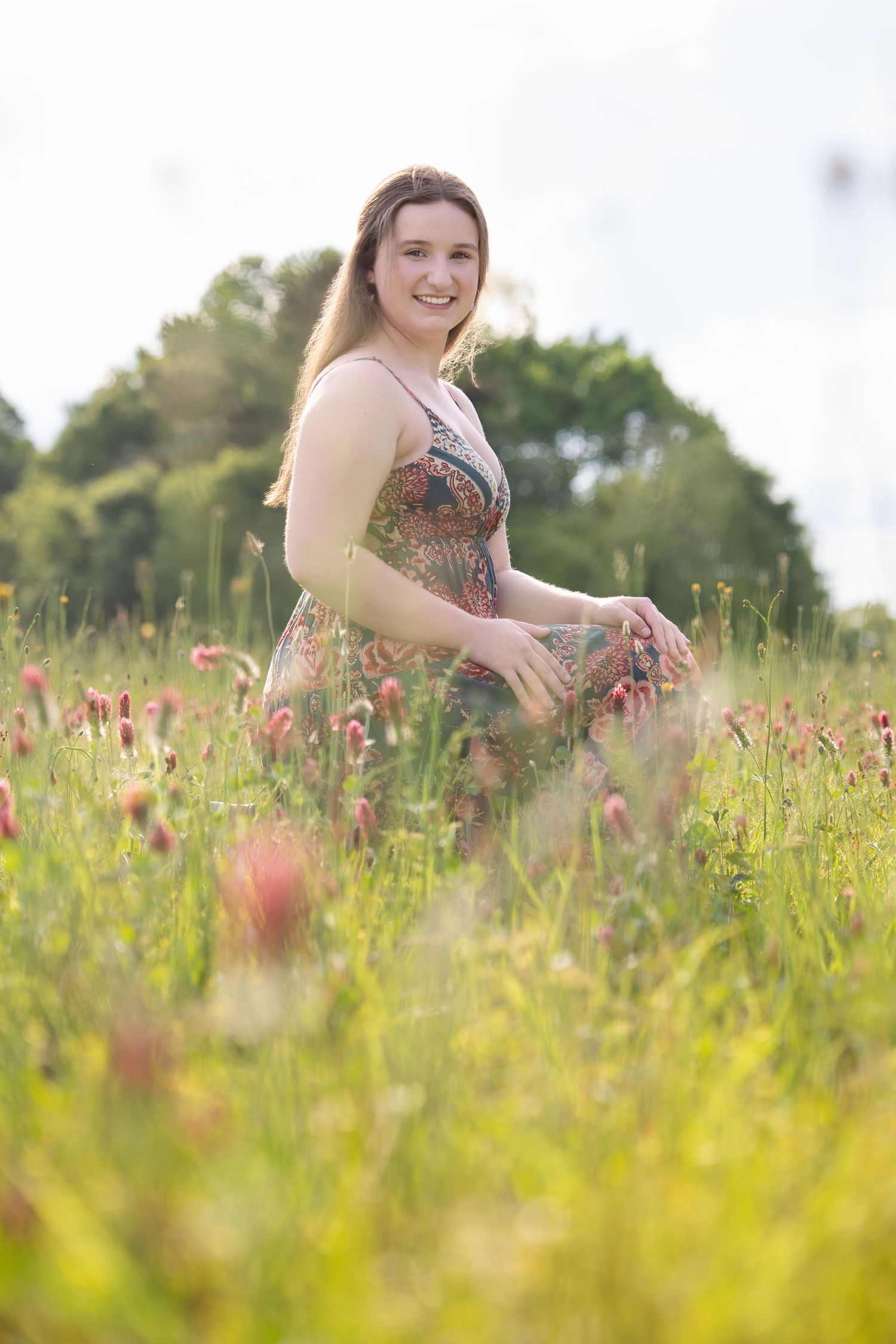 Person in a floral dress sits among tall wildflowers and grass in a sunny meadow on a summer day.