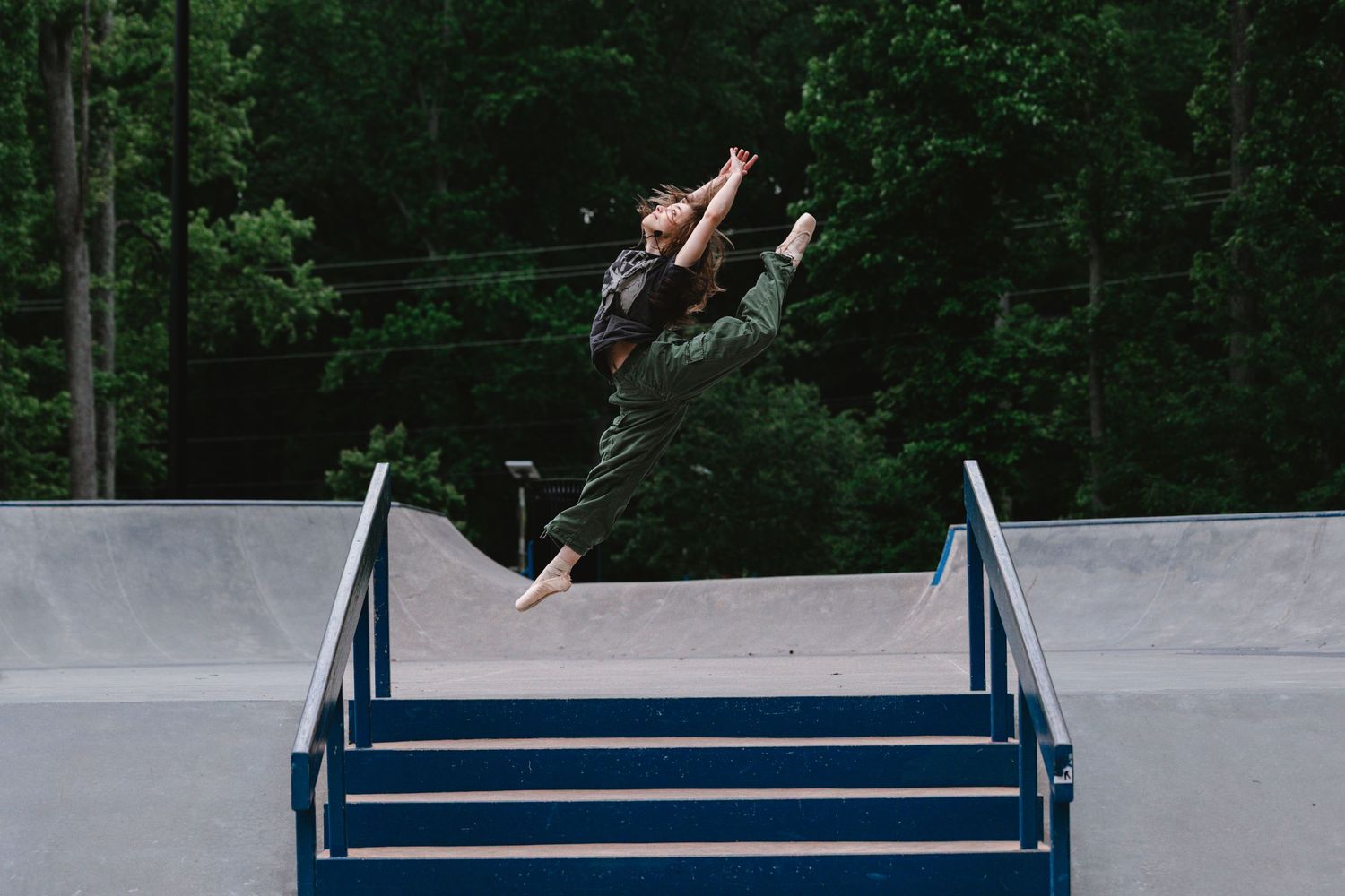 Skateboarder performs an aerial trick over a blue metal rail at a concrete skate park in a wooded setting.