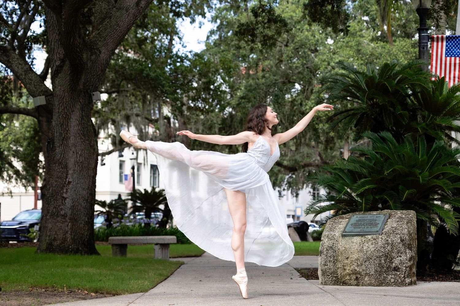 A dancer in a flowing white dress performs graceful movements in a lush outdoor garden setting with Spanish moss.