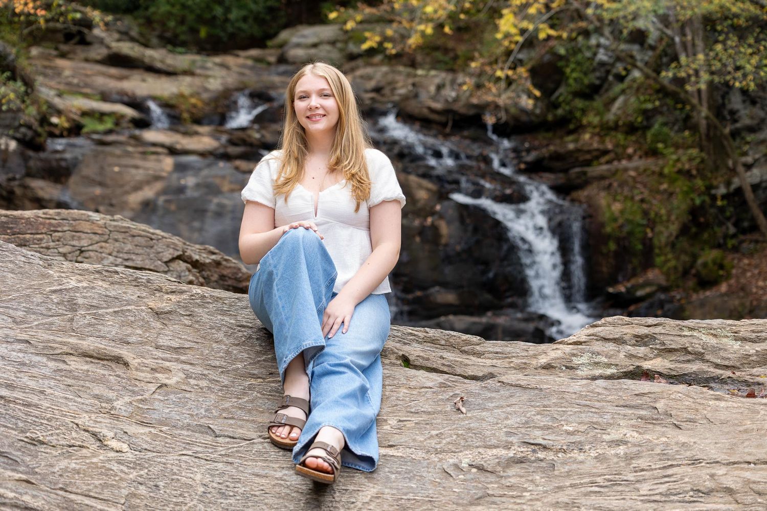 Person in light blue dress sits on large rock formation near cascading waterfall in scenic forest setting.