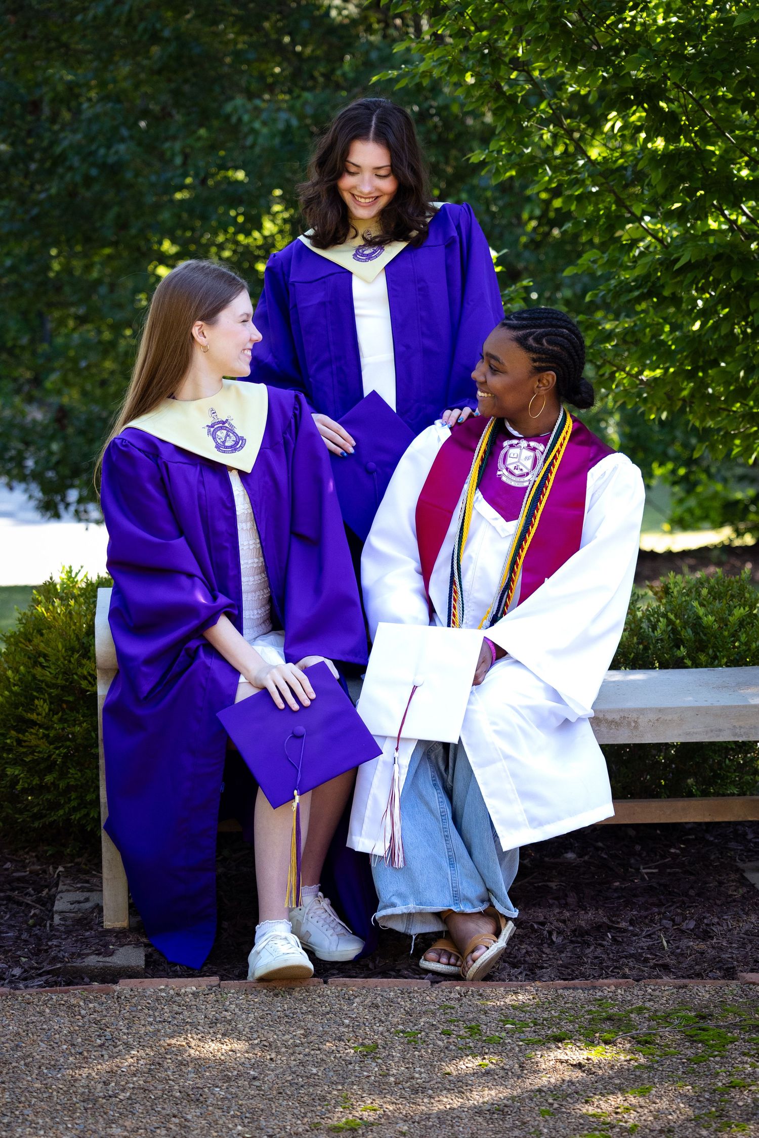 Three graduates in purple and white robes smile together outdoors on a bench, celebrating their academic achievement.