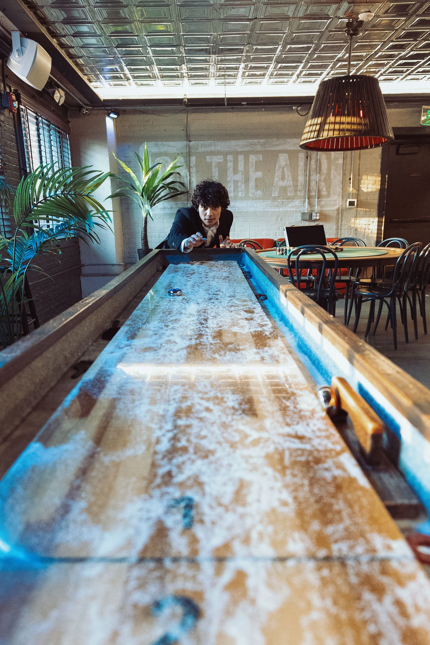 Man leaning over a shuffleboard table in a modern bar with pendant lighting and green plants.