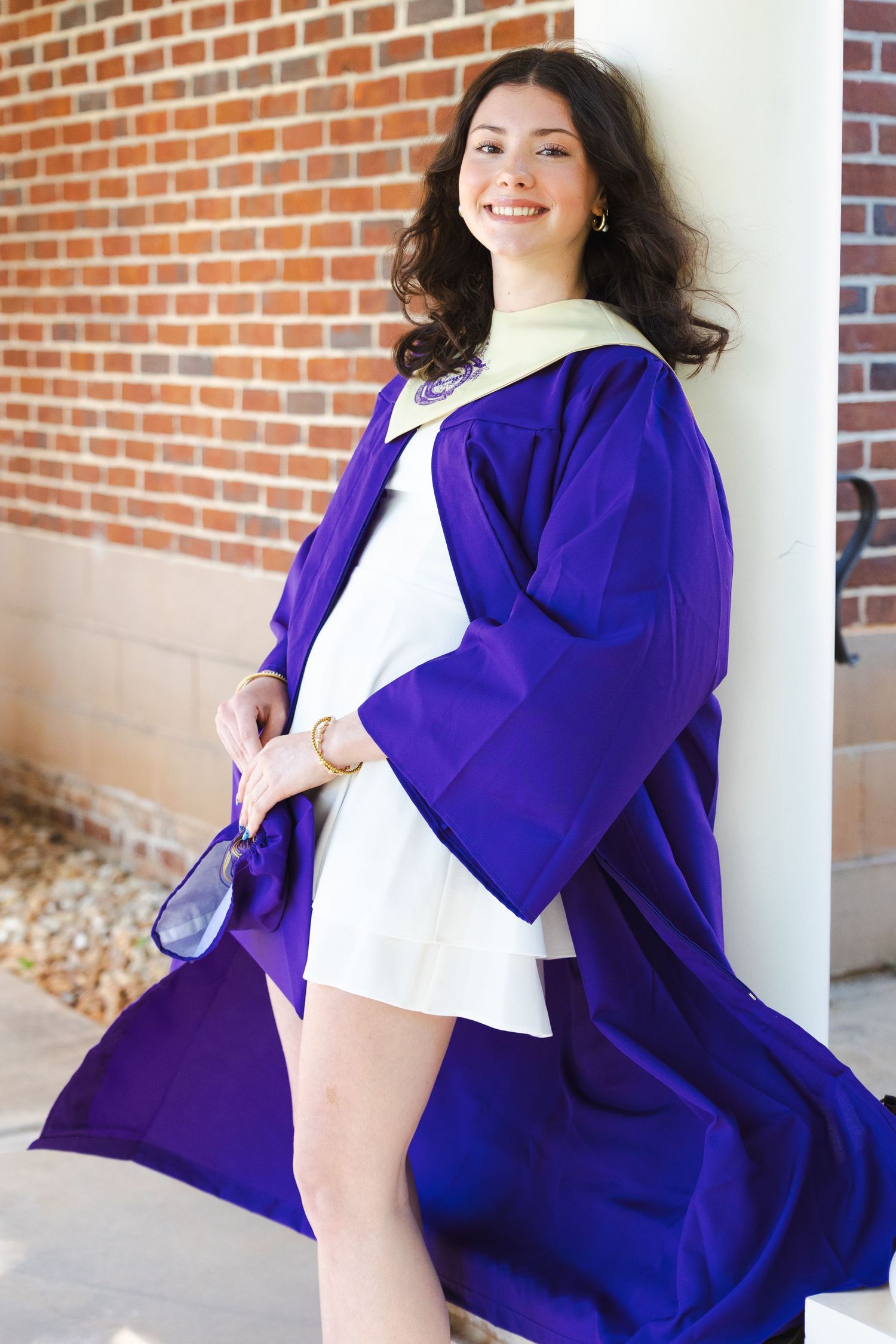 Smiling young woman in purple graduation gown leaning against white pillar with brick wall background.