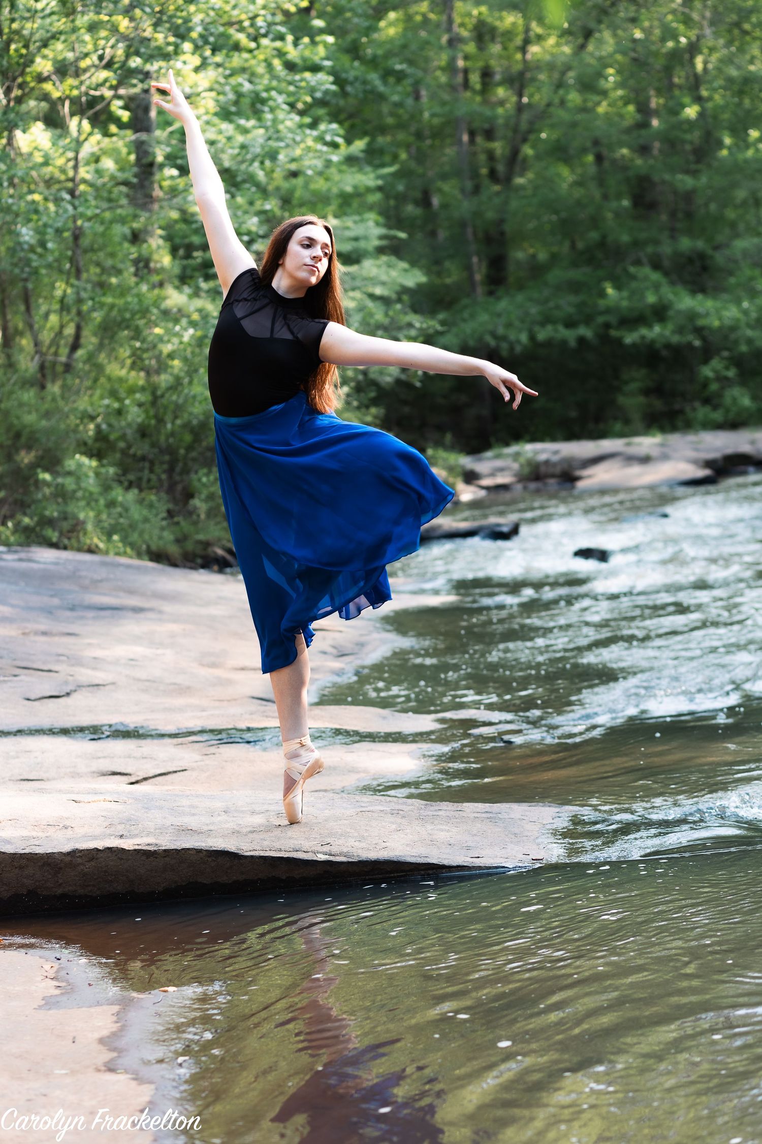 A dancer in a flowing blue dress performs graceful movements on rocks by a flowing stream in a serene forest setting.