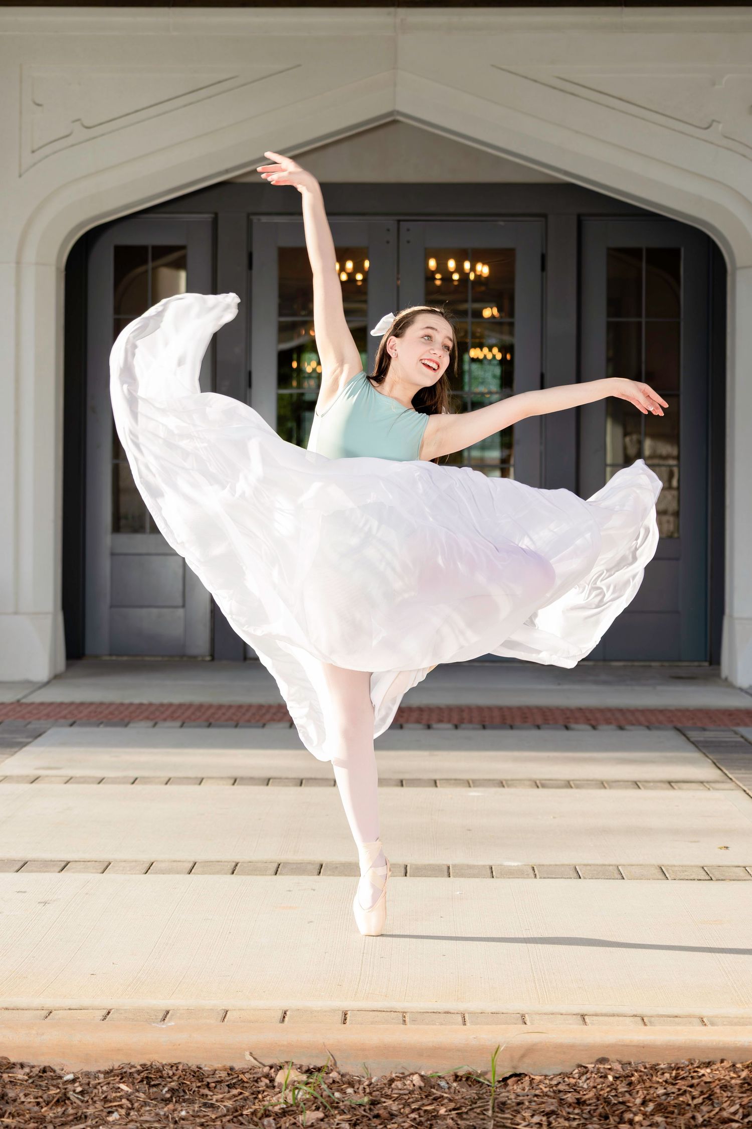 Dancer in flowing white dress performing elegant ballet moves on stone patio.