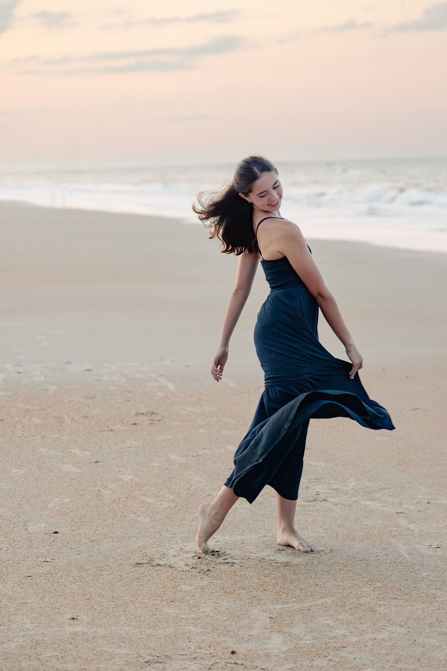 Person in navy dress twirling and dancing joyfully on empty beach at sunset.