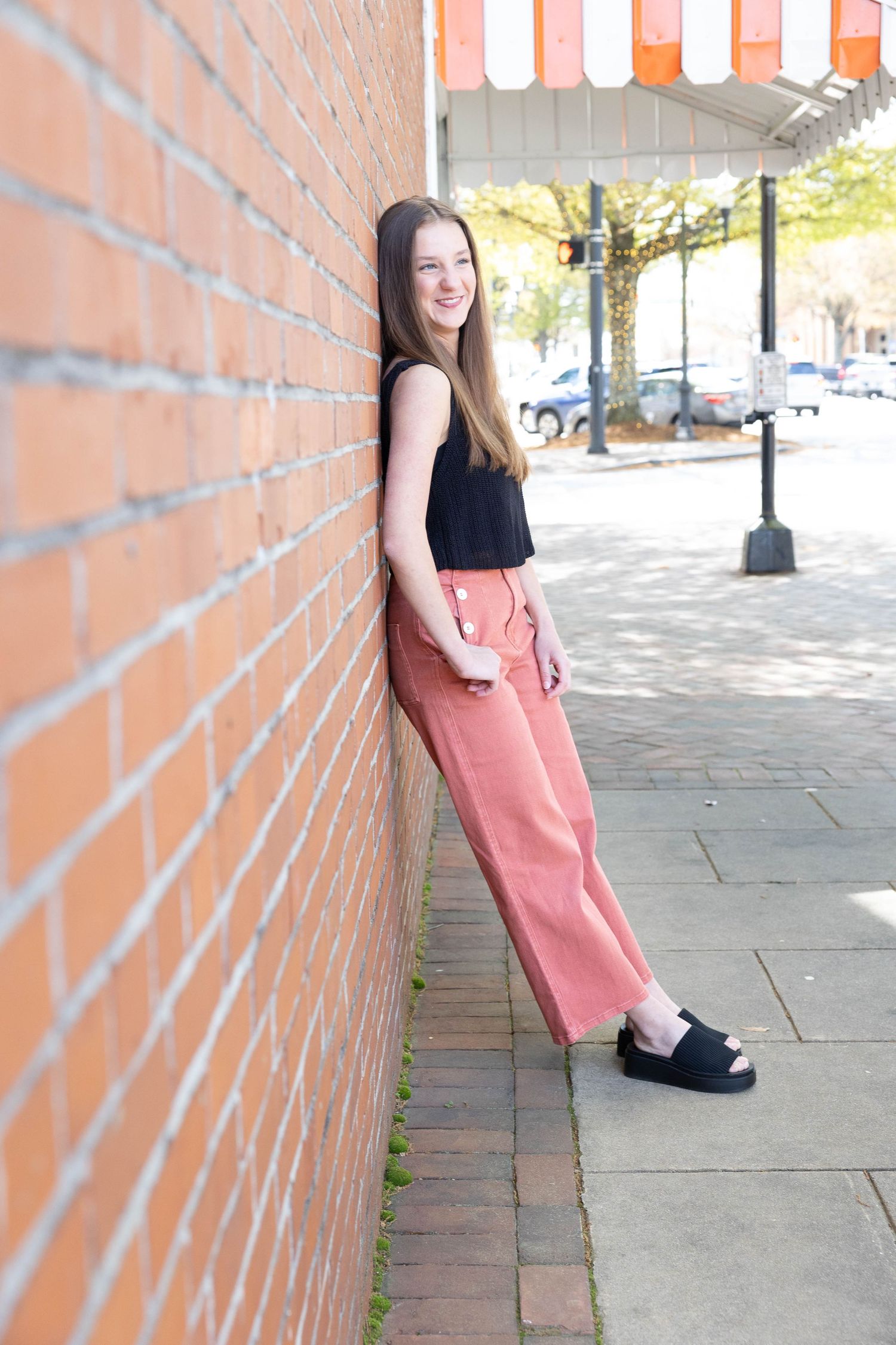 Person in a black top and pink skirt poses against a brick wall on a sunny day.