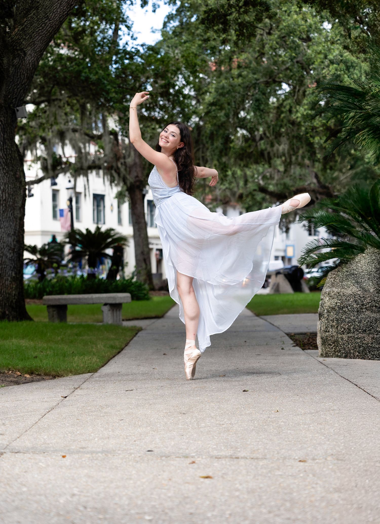 A dancer in white performs graceful ballet moves on a concrete path surrounded by trees.