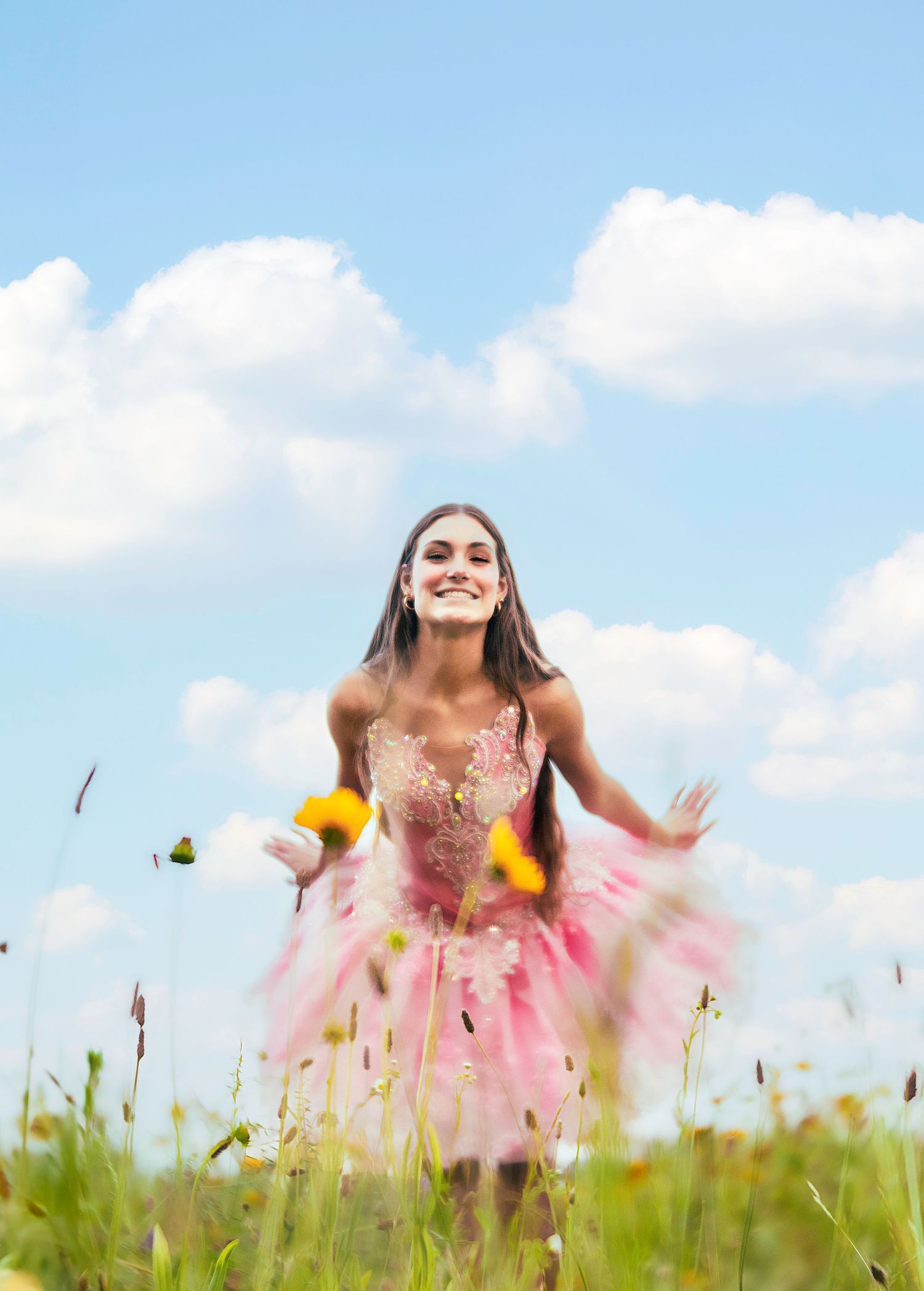Young woman in pink tulle dress twirling in a wildflower meadow under a bright blue sky with fluffy clouds.